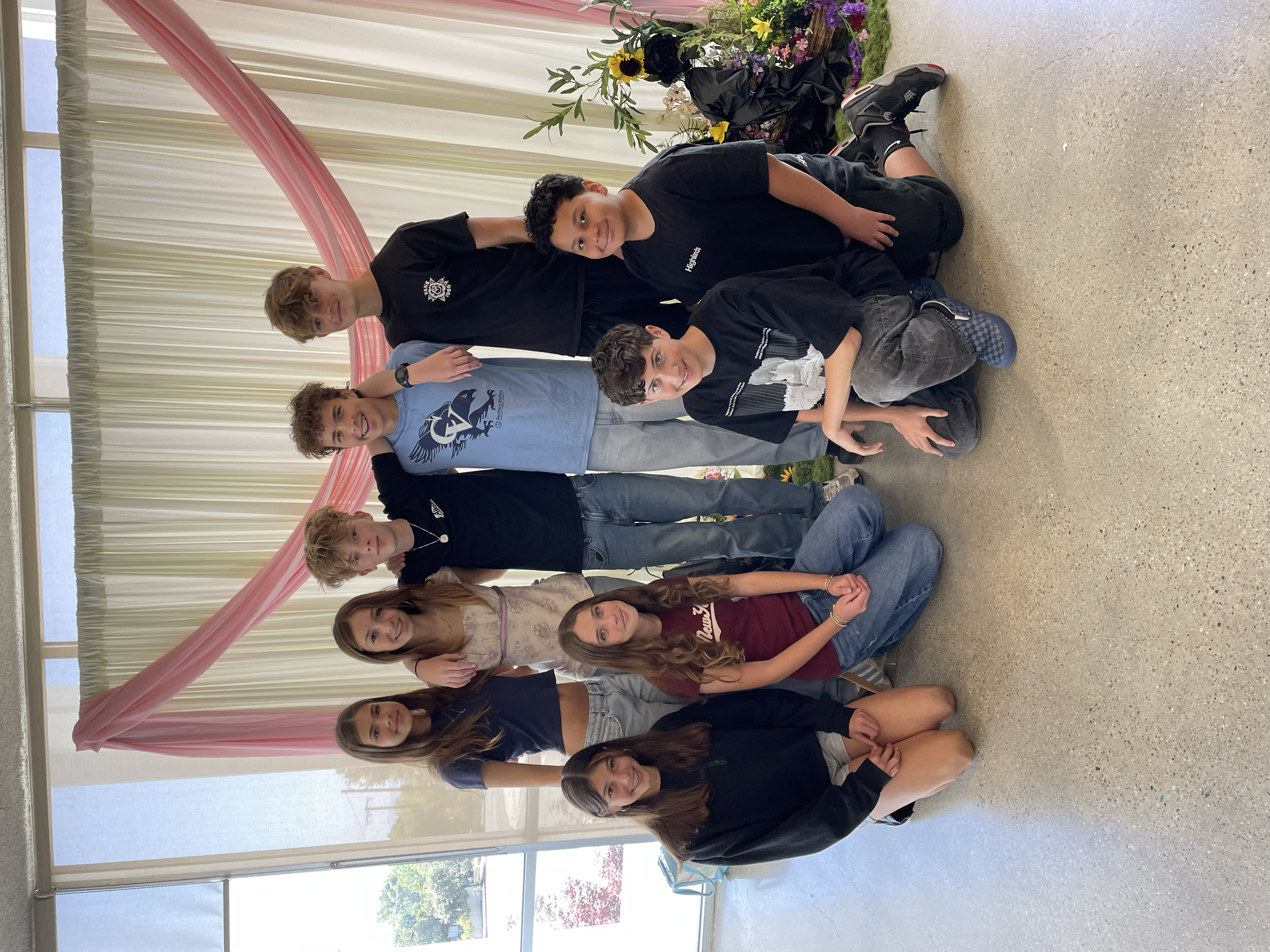 A group of young people, mostly teenagers, are posing indoors in front of large windows with white curtains. They are sitting or kneeling on the floor, with some smiling at the camera. Behind them, there are pink and white decorative drapes and a flo
