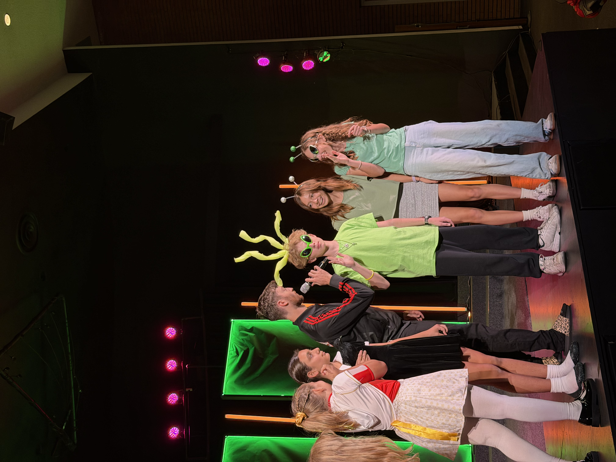 Group of kids dressed in futuristic and colorful costumes standing on a stage with colorful lights and a dark background.