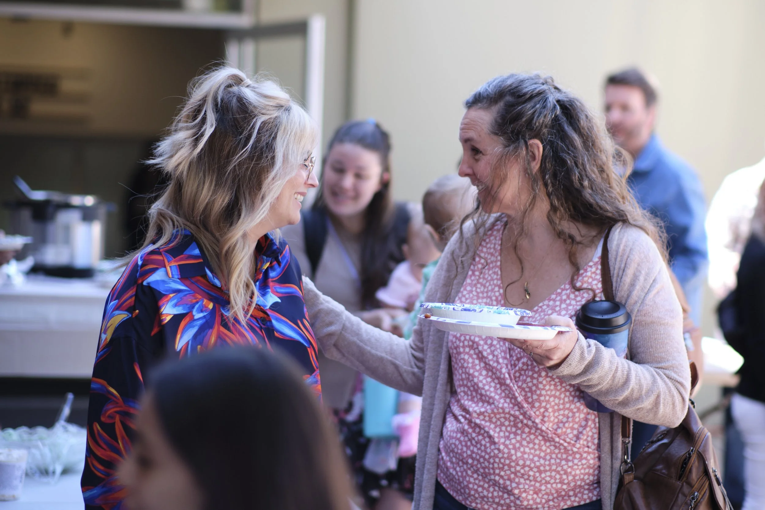 Two women are smiling at each other during an outdoor event. One woman is handing a food tray to the other. Several people are in the background, and there is a coffee pot on a table.