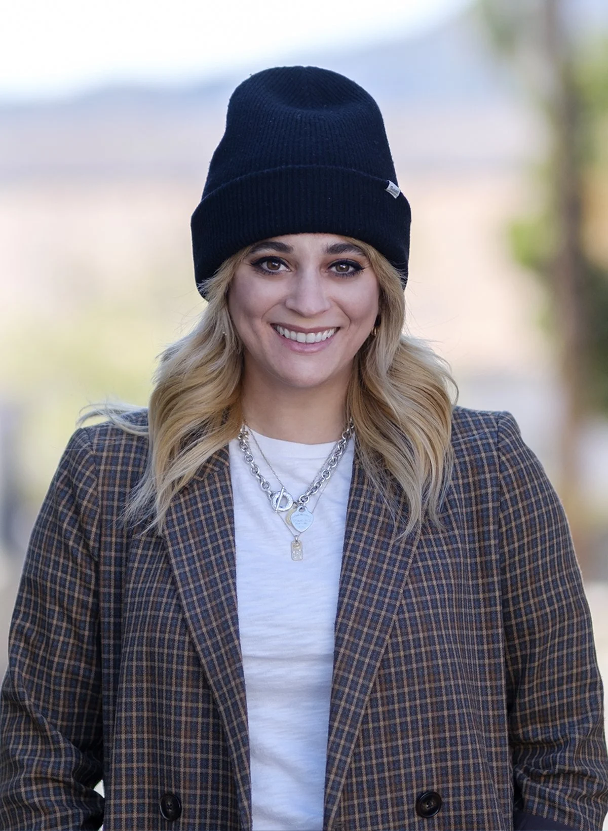 A woman with blonde hair smiling, wearing a black beanie, layered necklaces, a white t-shirt, and a brown checkered blazer, outdoors with a blurred background.