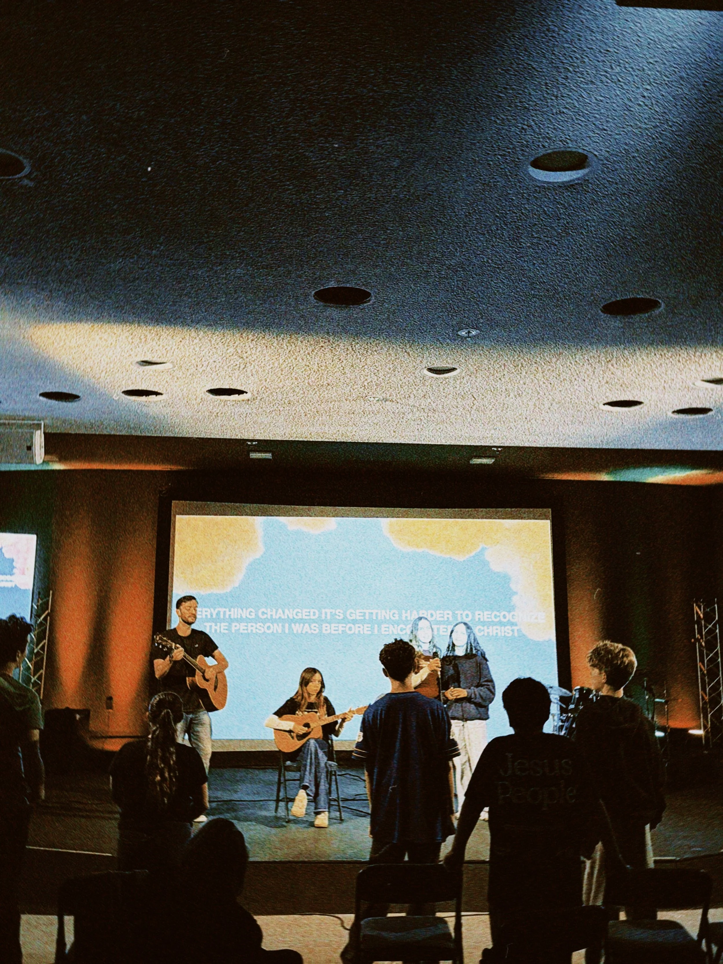 People gathered in a room watching a presentation on a large screen, some are playing guitars, and a few are standing while others are seated or standing around the room.