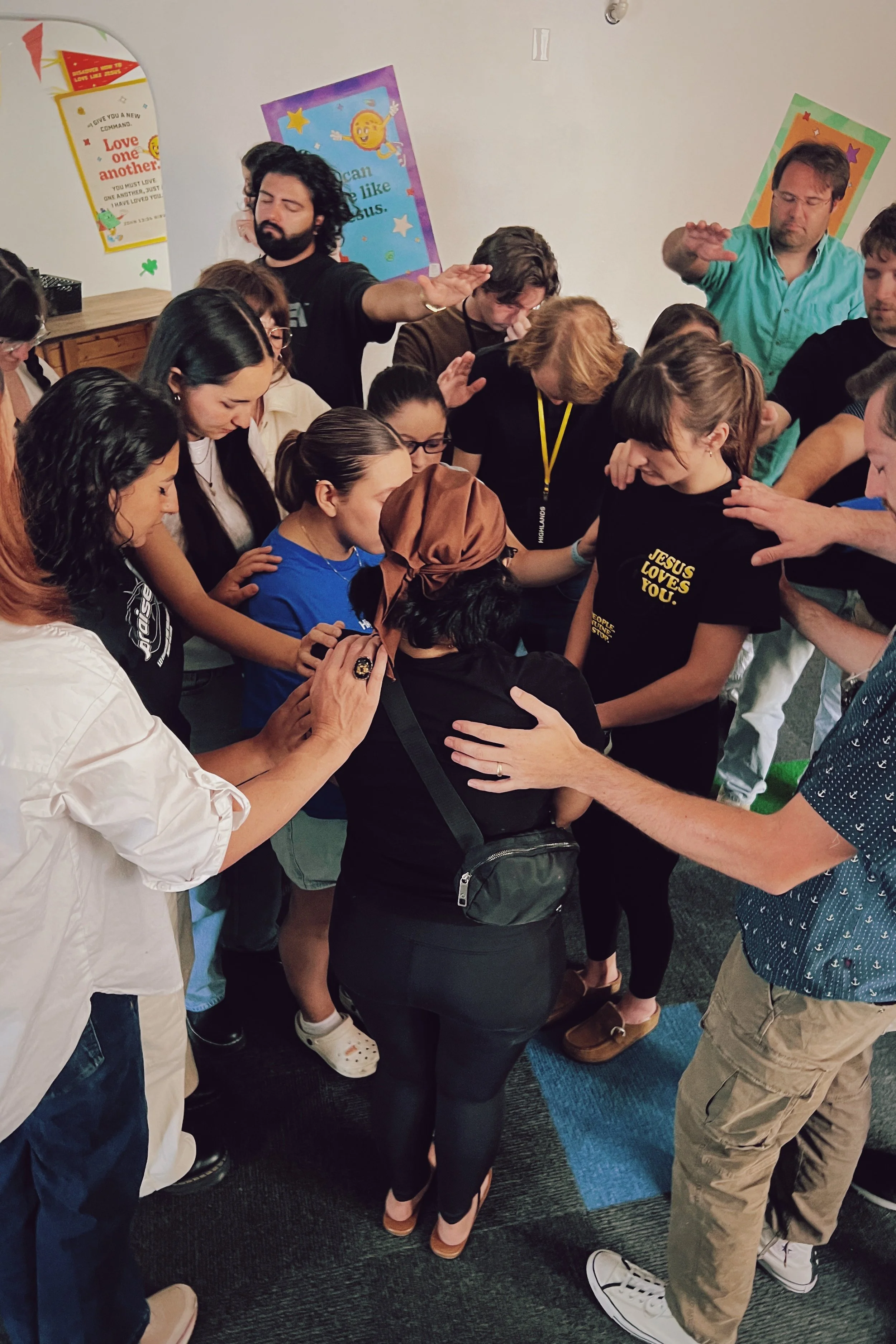 A diverse group of people are gathered in a circle with their hands joined in prayer or meditation, leaning their heads in with closed eyes, in an indoor setting.
