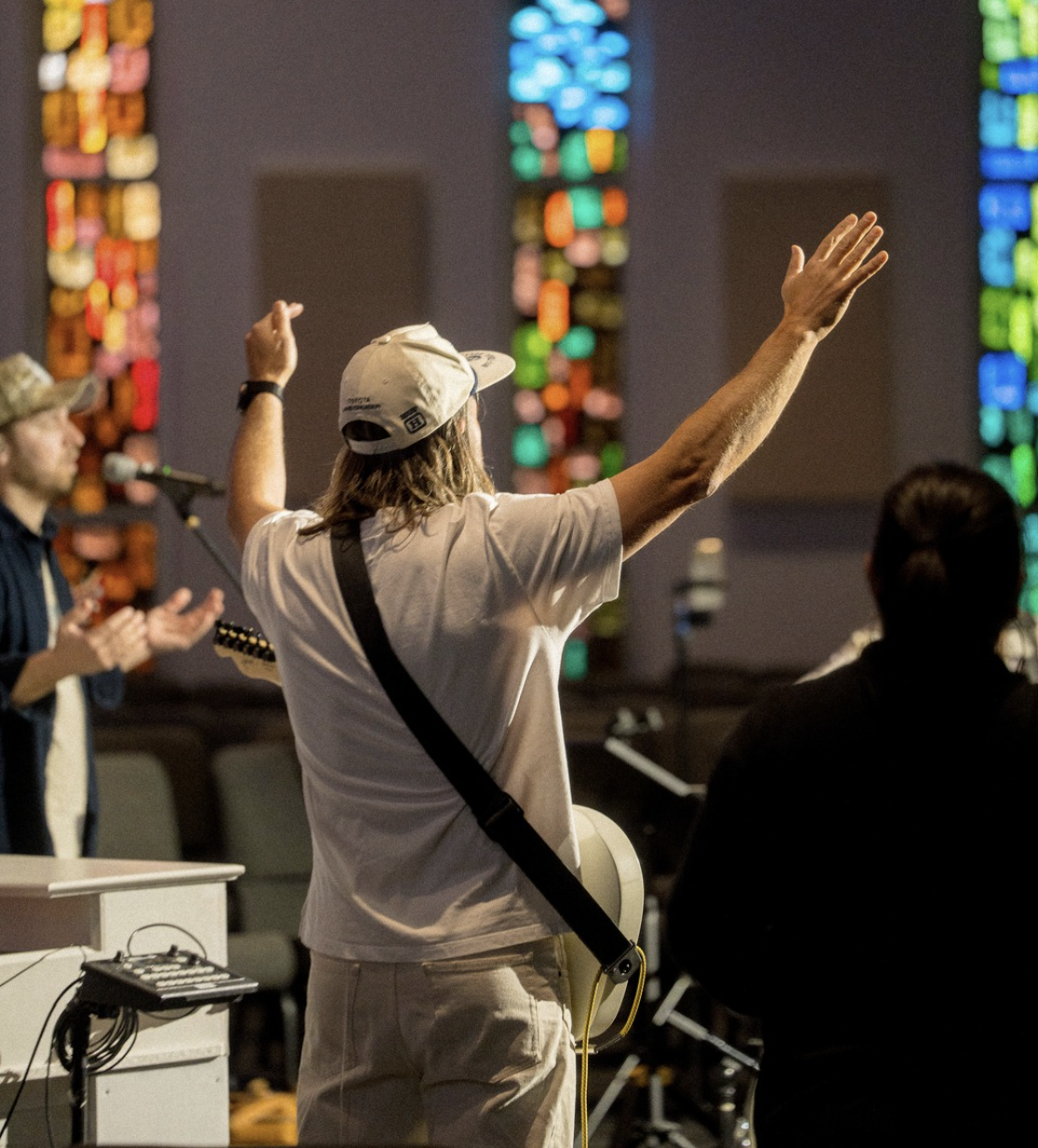 A person wearing a cap and a white t-shirt raising their arms during a musical performance in a church or auditorium with stained glass windows.