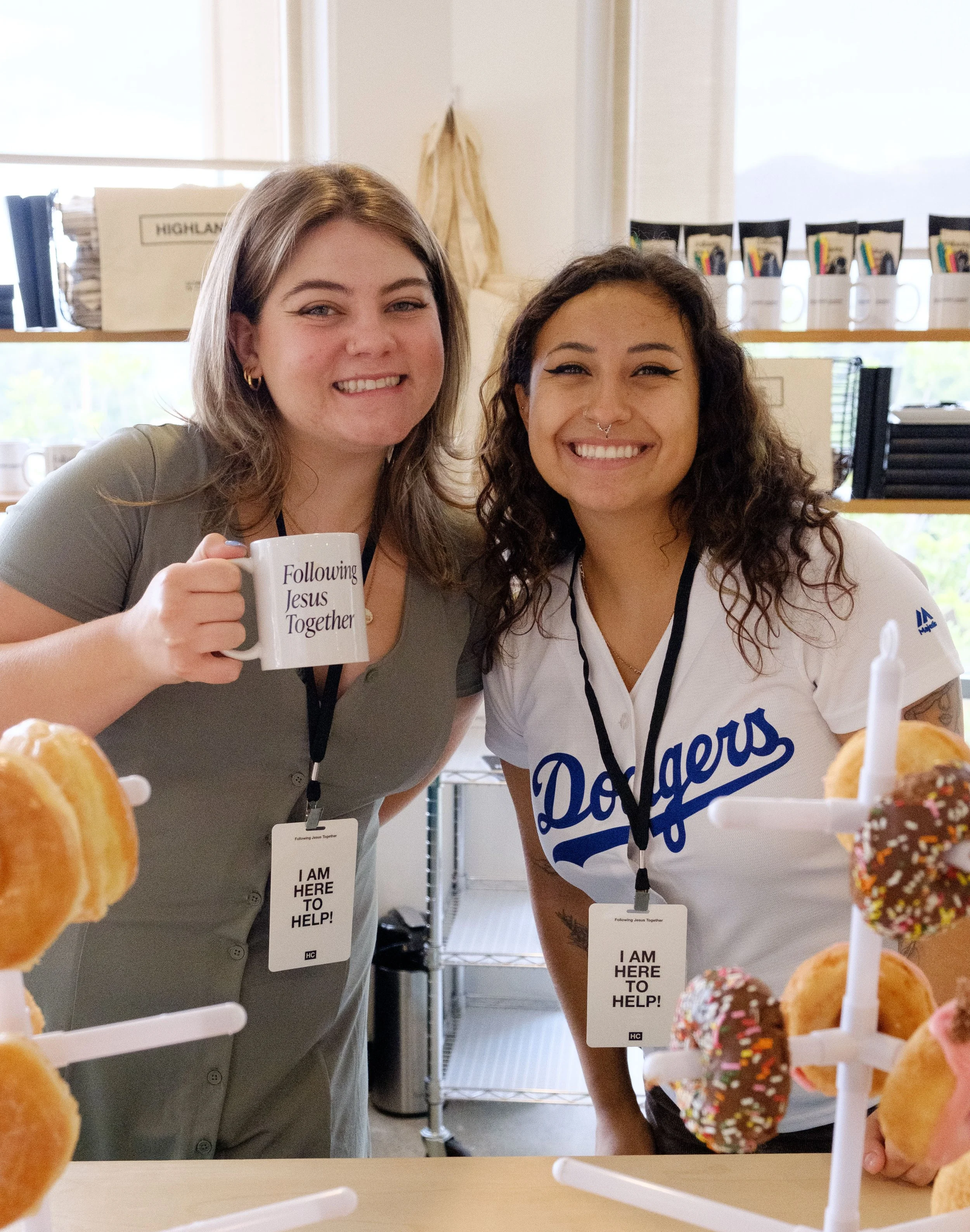 Two women smile at the camera behind a table of donuts, one holding a mug that says "Following Jesus Together" and both wearing lanyards with badges that say "I am here to help." Behind them are shelves with books and mugs, a window, and a trash can.
