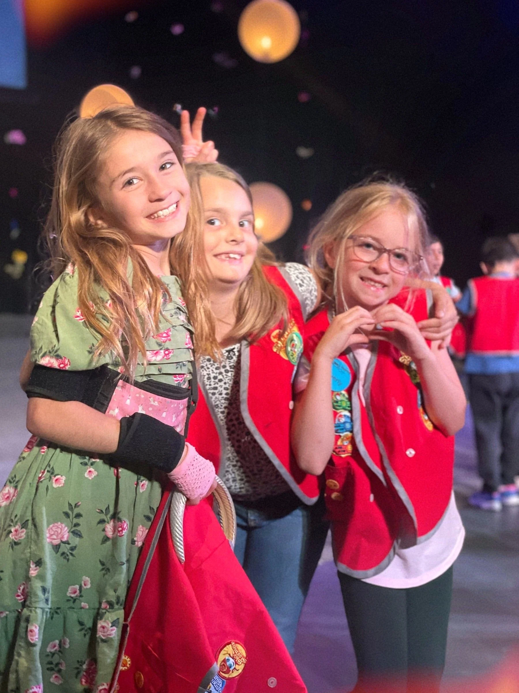 Three young girls standing together smiling at a party or celebration, with a dark background and floating decorative lights.