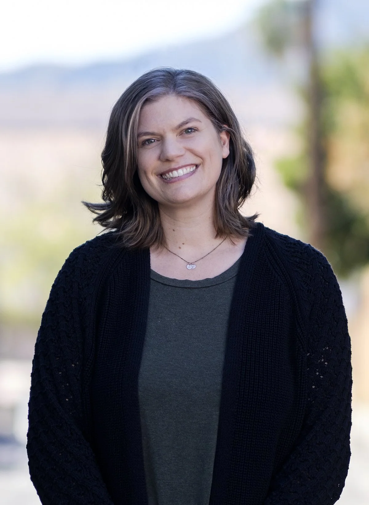 A smiling woman with shoulder-length brown hair and fair skin, wearing a dark green shirt, a black knit cardigan, and a silver necklace, standing outdoors with a blurred background of trees and sky.