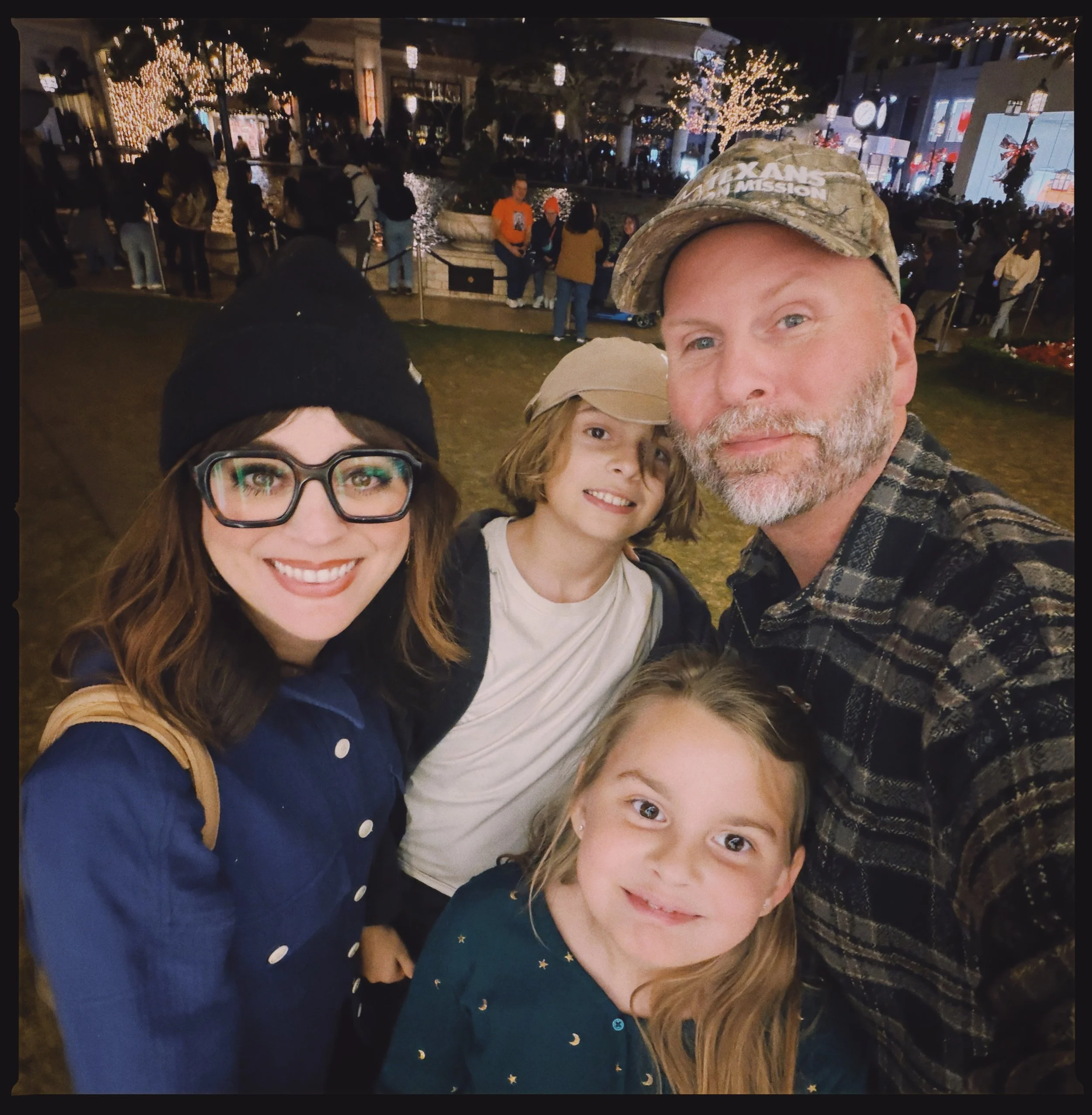 Family selfie in a crowded outdoor area with festive lights, trees wrapped in Christmas lights, and storefronts decorated for the holidays. The group includes two women and one young girl smiling at the camera.