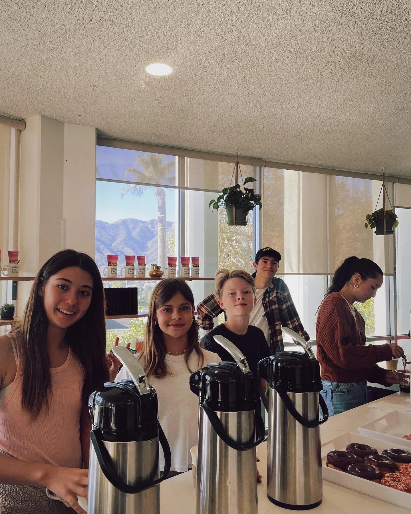Group of five young people standing behind a counter with coffee dispensers and a tray of donuts, inside a bright room with large windows showing mountains and palm trees outside.