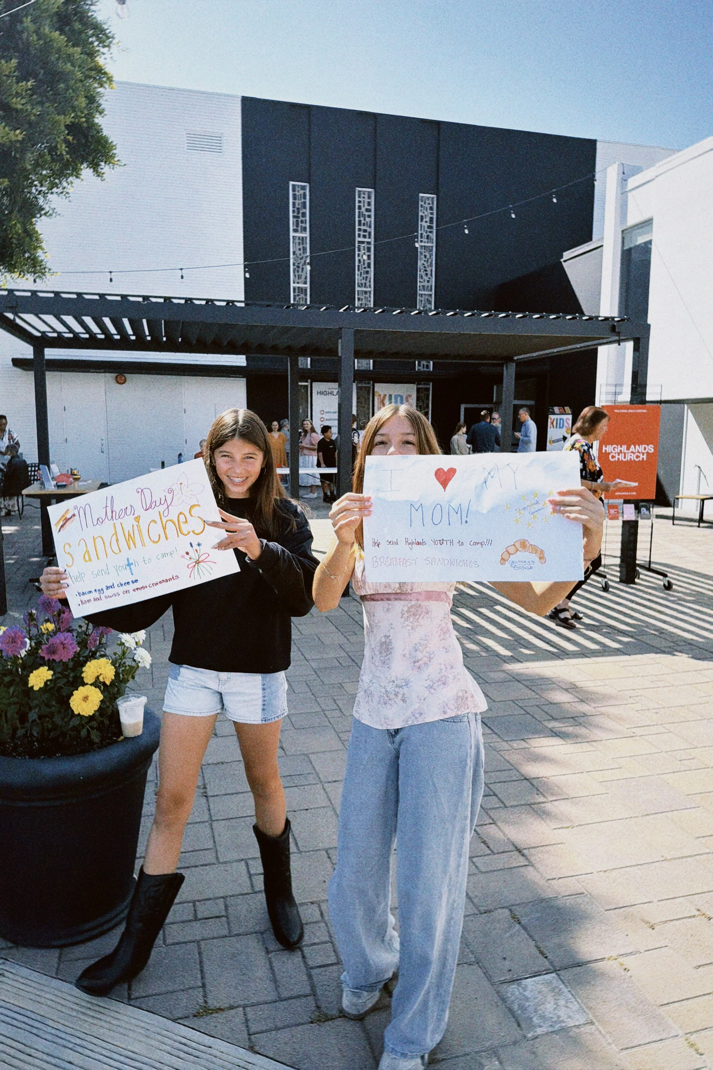 Two young women holding handmade signs supporting their mom at a community event outside a modern building. One sign reads 'Mother Day Sandwiches' and the other 'I ❤️ MY MOM!'. There are other people in the background near a table and a sign for High