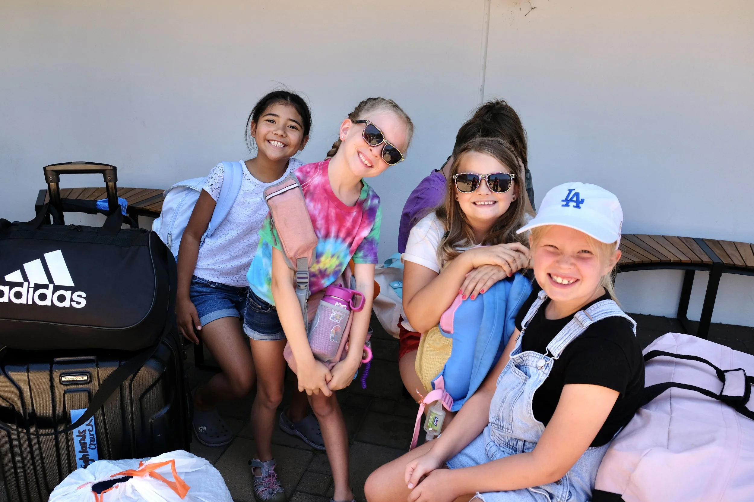Group of smiling children with backpacks and sunglasses, standing on a bench, with luggage nearby, against a plain wall.