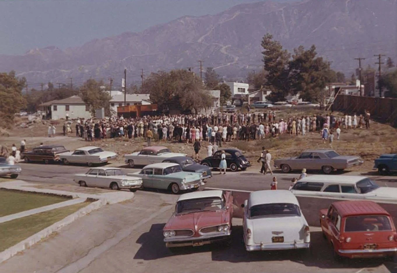 A large gathering of people outdoors in a grassy area, with cars parked in the foreground on a parking lot and street, during daytime. The crowd appears to be attending an event or assembly with mountains in the background.