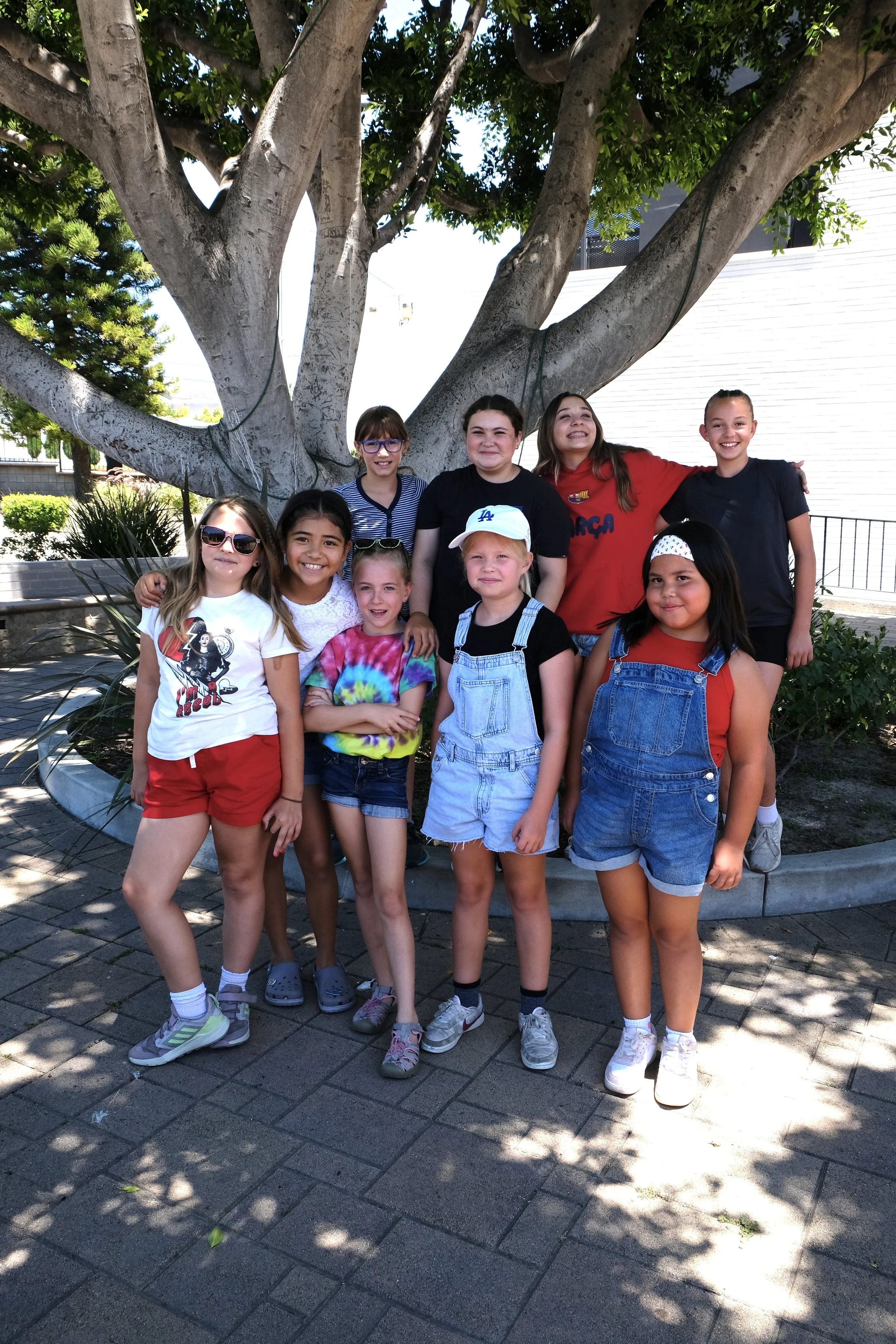 A group of nine diverse children, smiling and standing in front of a large tree in an outdoor setting. Some kids are wearing sunglasses, colorful clothes, and casual shoes.