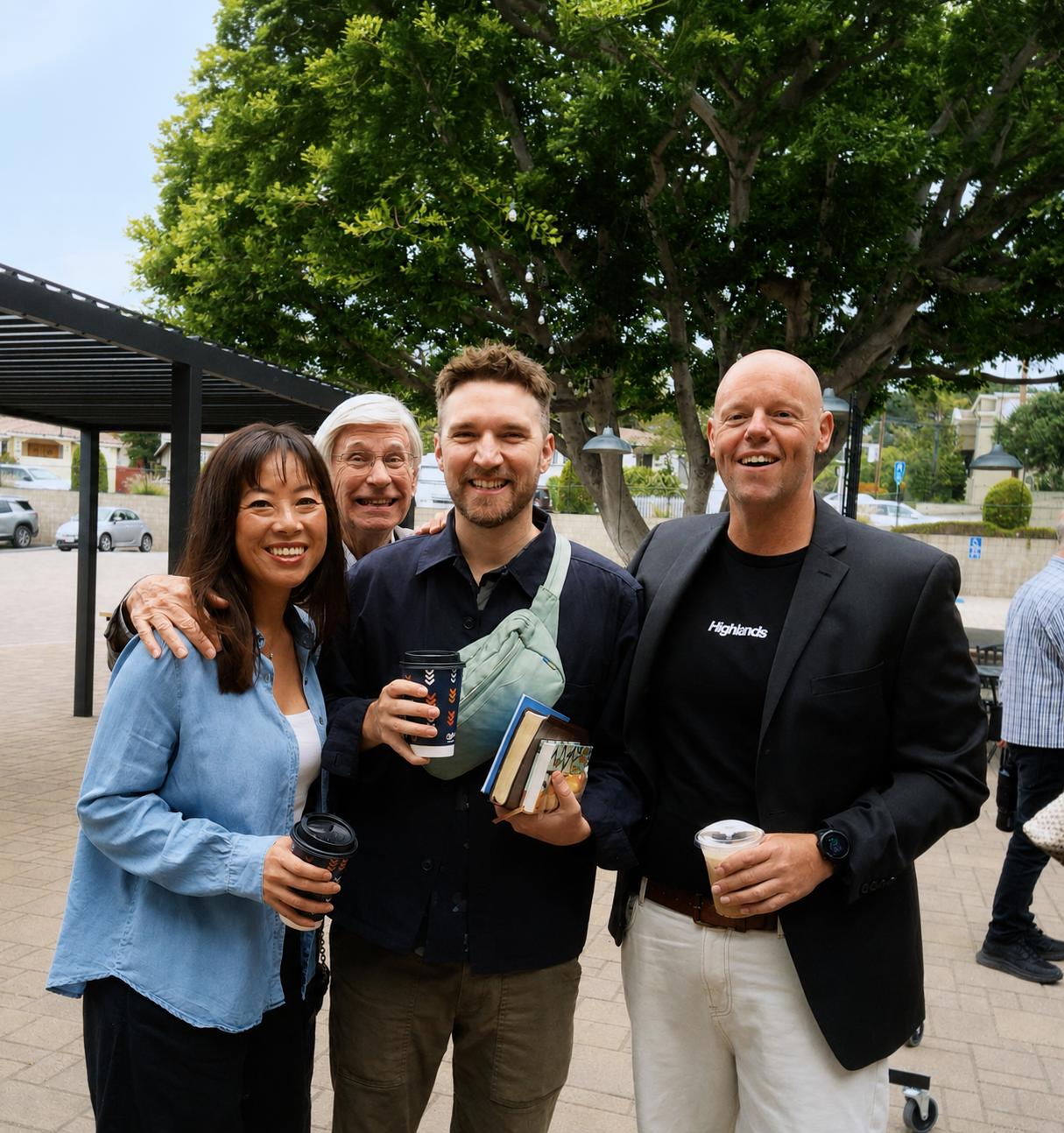 Group of four diverse adults smiling outdoors, holding coffee cups and books, standing under a tree with a park setting in the background.