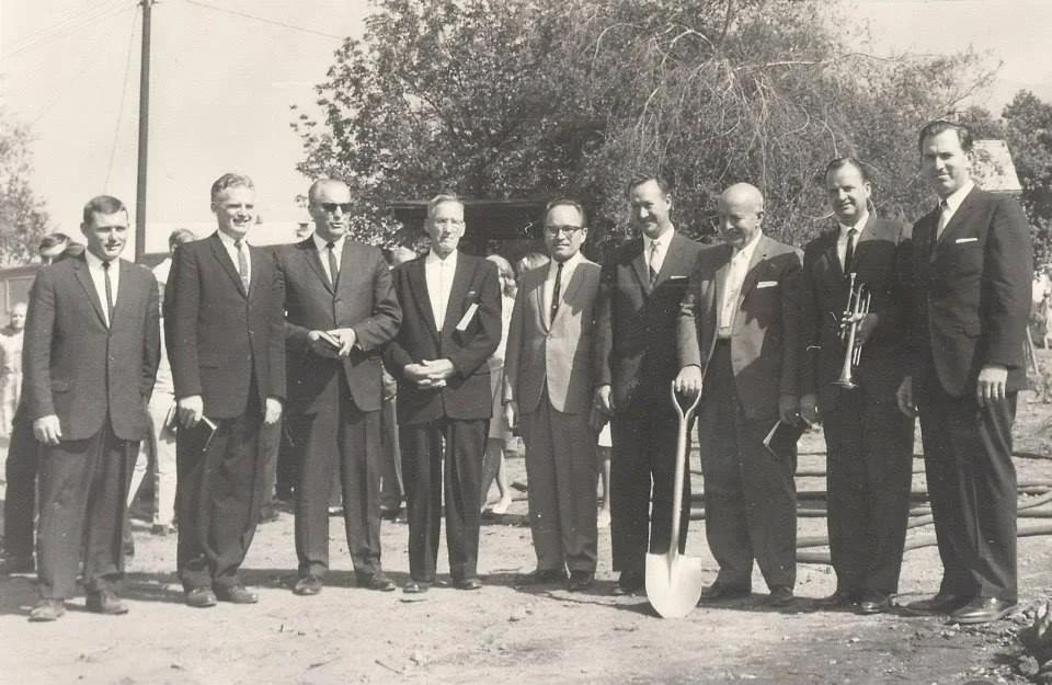 Black and white photo of nine men in suits standing outdoors, holding a shovel and a trumpet, with trees and other people in the background.