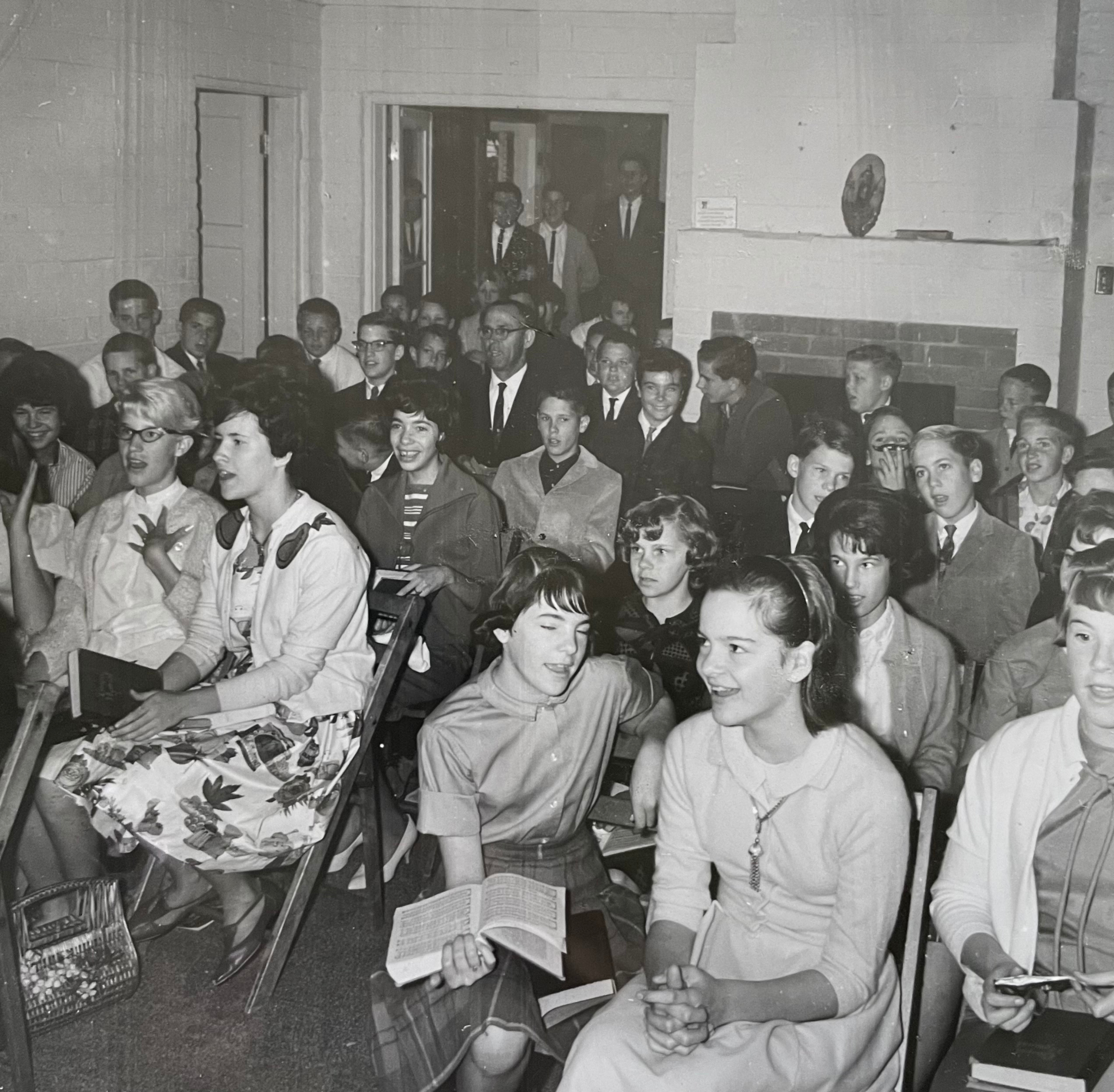 Black and white photograph of a crowded indoor scene with many people, mostly young, engaged in various activities. A few individuals appear to be singing or reciting, and a woman is holding a large opened book or magazine while sitting on the floor. The room has brick walls, a fireplace, and shelves with books and decorations.