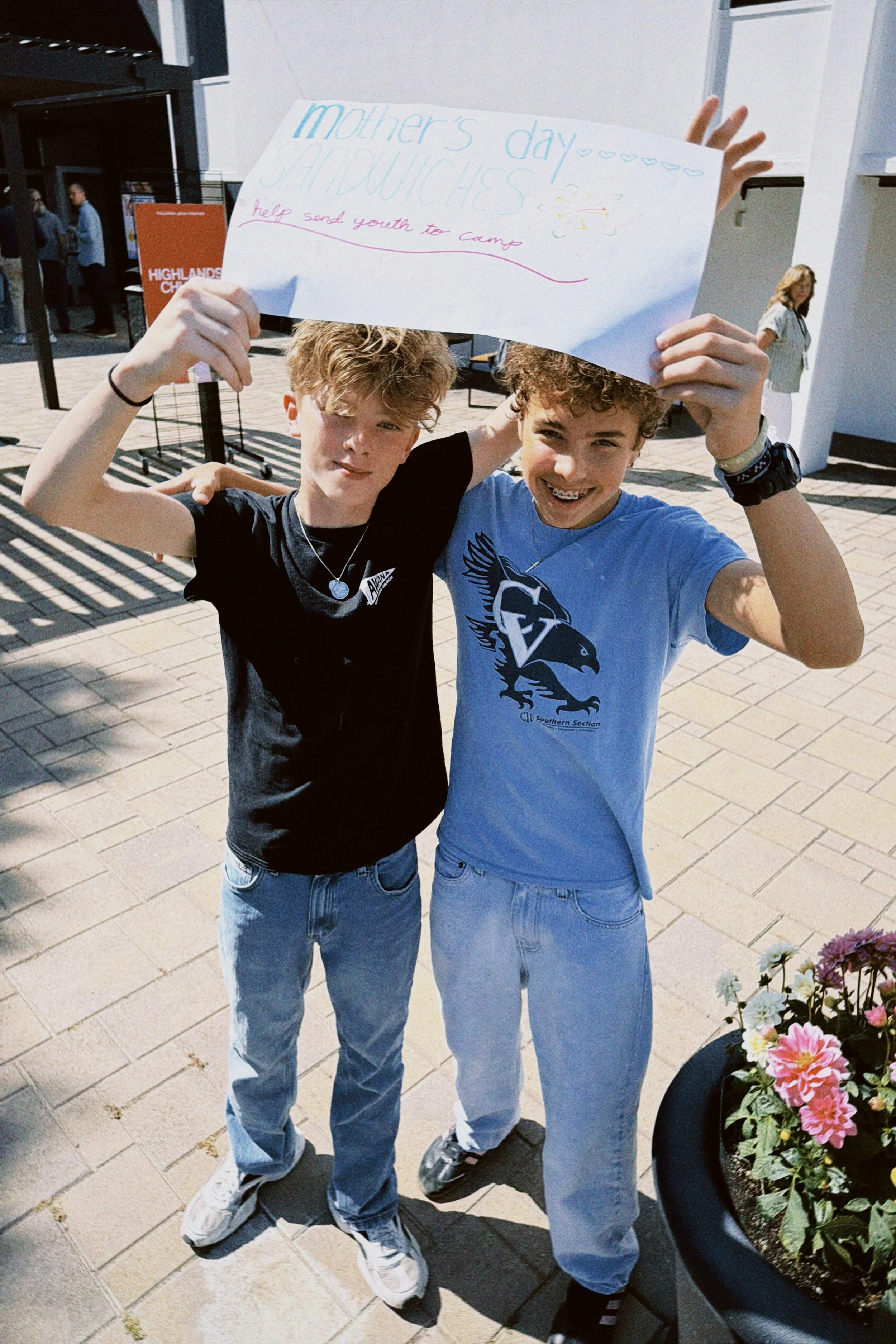 Two young males standing outside, smiling, holding a handmade sign that reads "Mother's Day Shadows" with additional text about helping send youth to camp. They are wearing casual clothing, jeans, and sneakers, with one wearing a wristwatch and the o