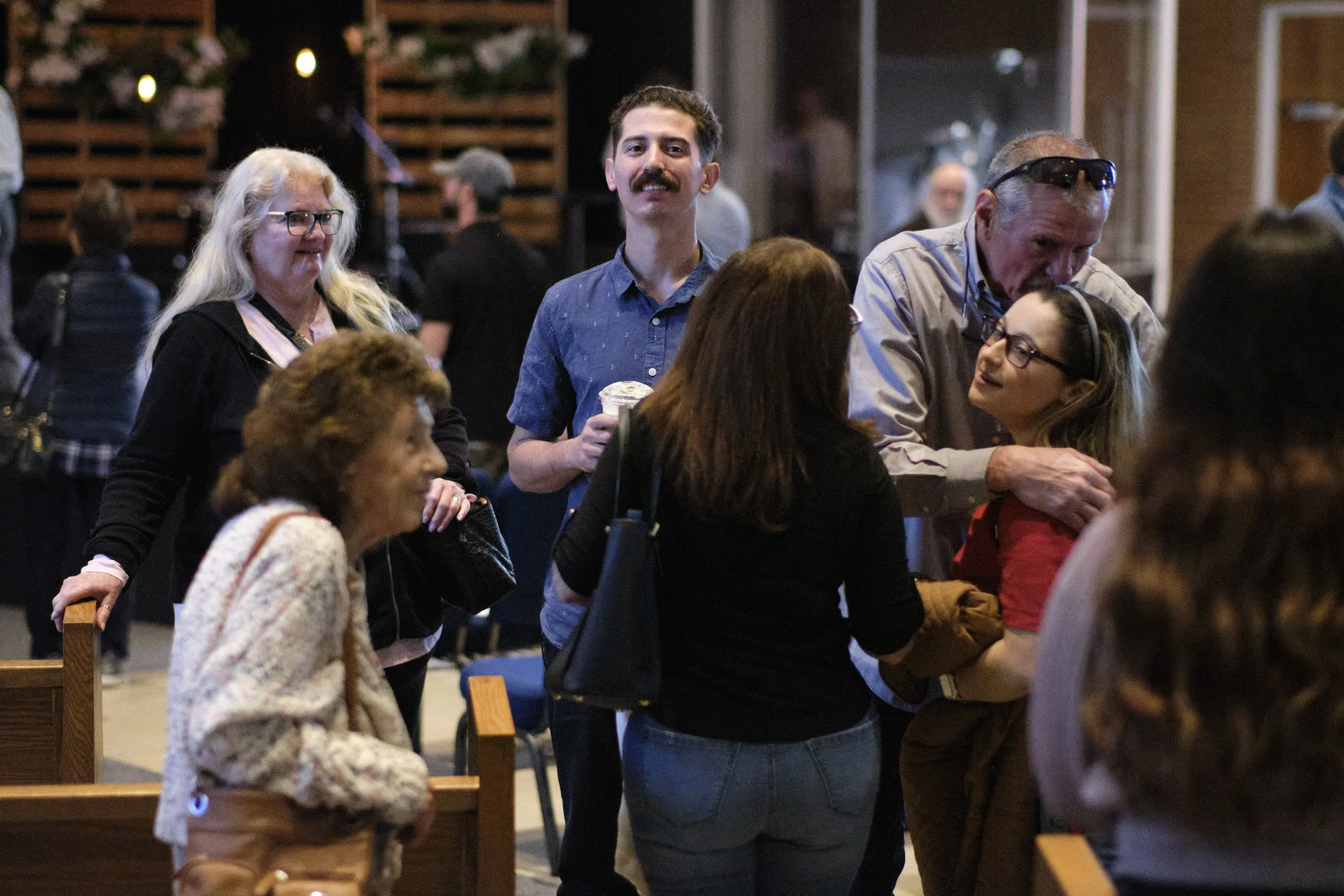 Group of people socializing indoors, some standing and some sitting, with a man in a blue shirt and mustache holding a drink in center.