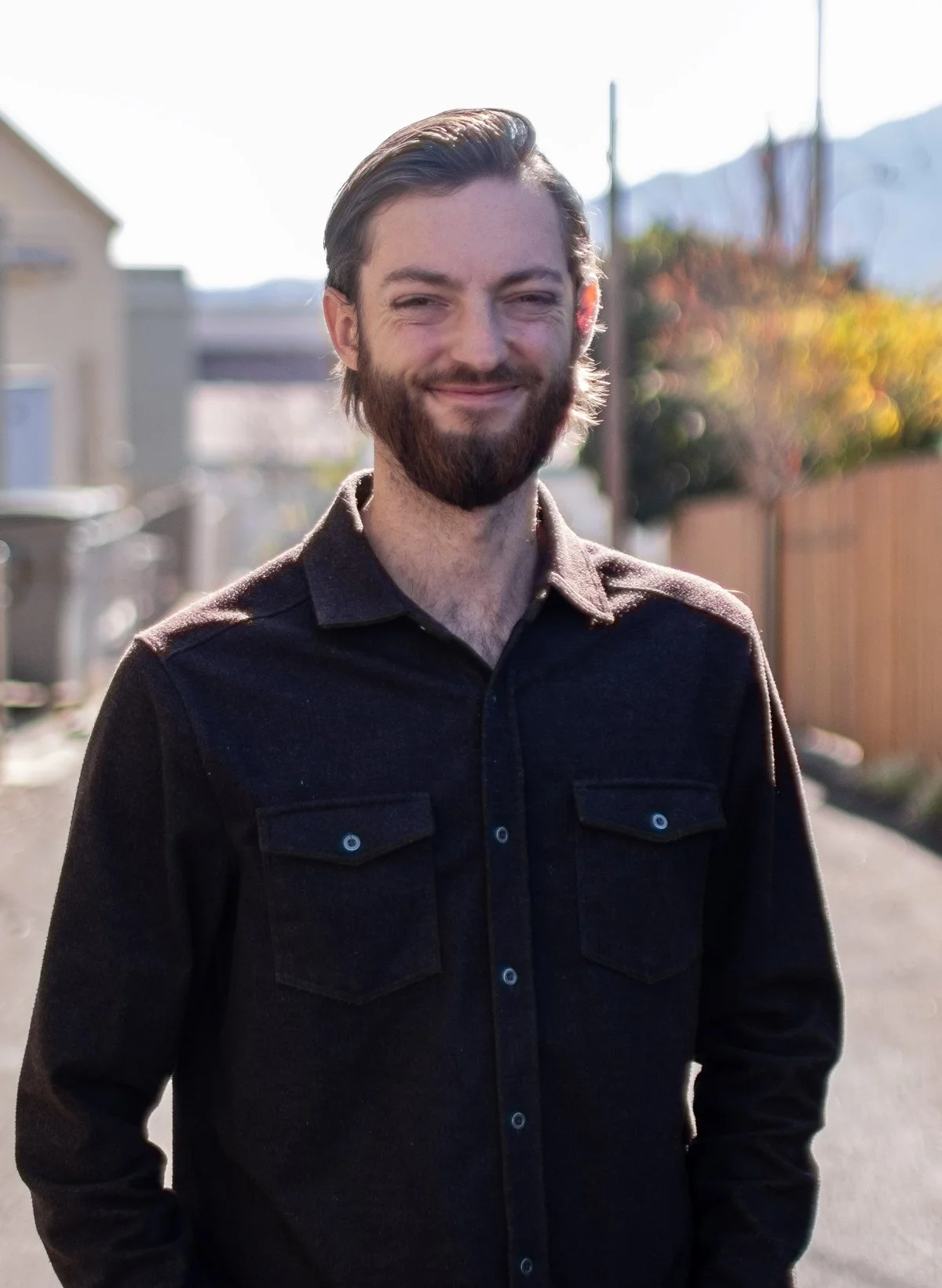 A smiling man with a beard stands outdoors in bright sunlight with houses, trees, and mountains in the background.