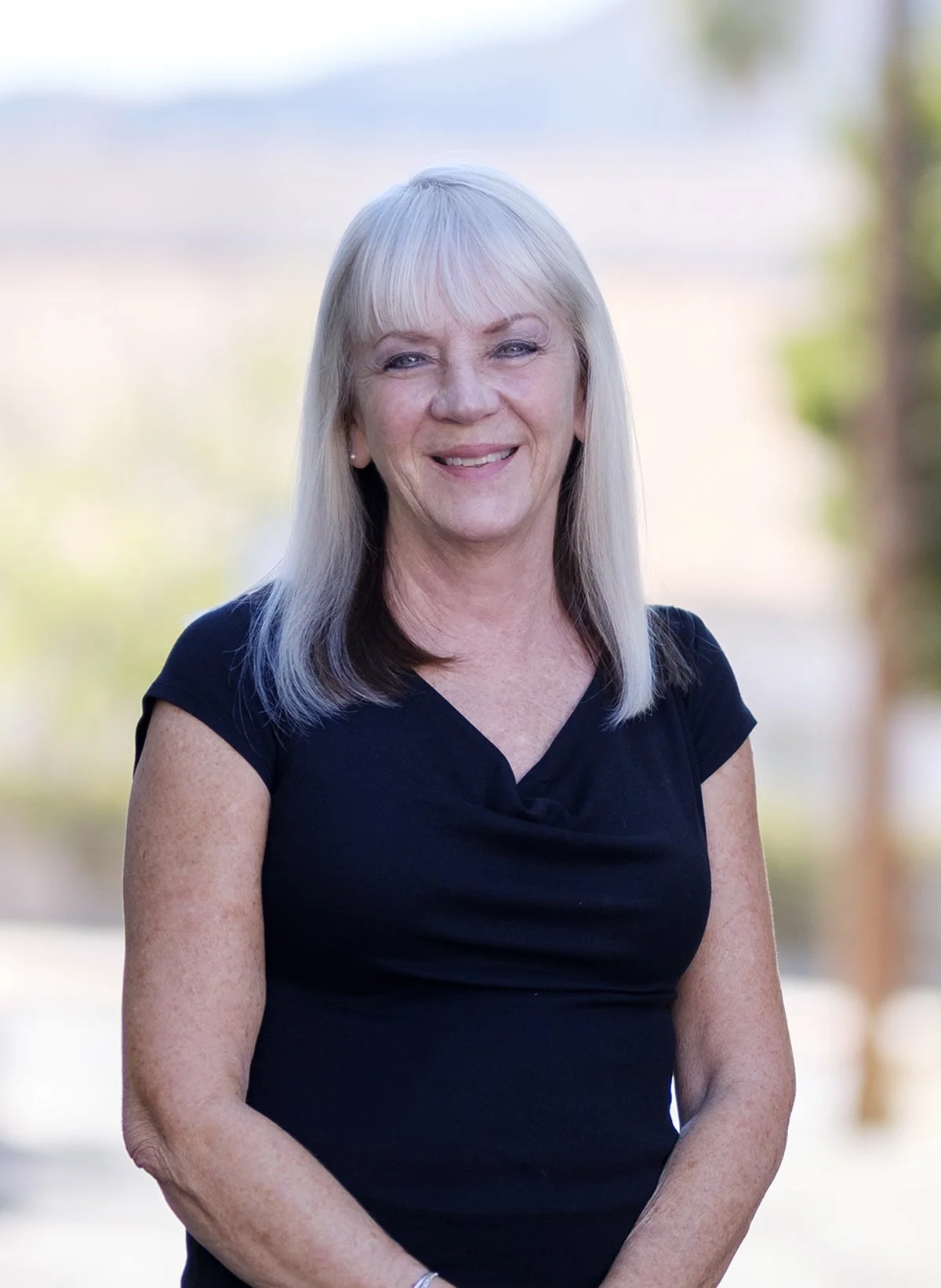 A middle-aged woman with long, straight, platinum blonde hair, smiling outdoors with a blurred background of trees and sky, wearing a black short-sleeved top.