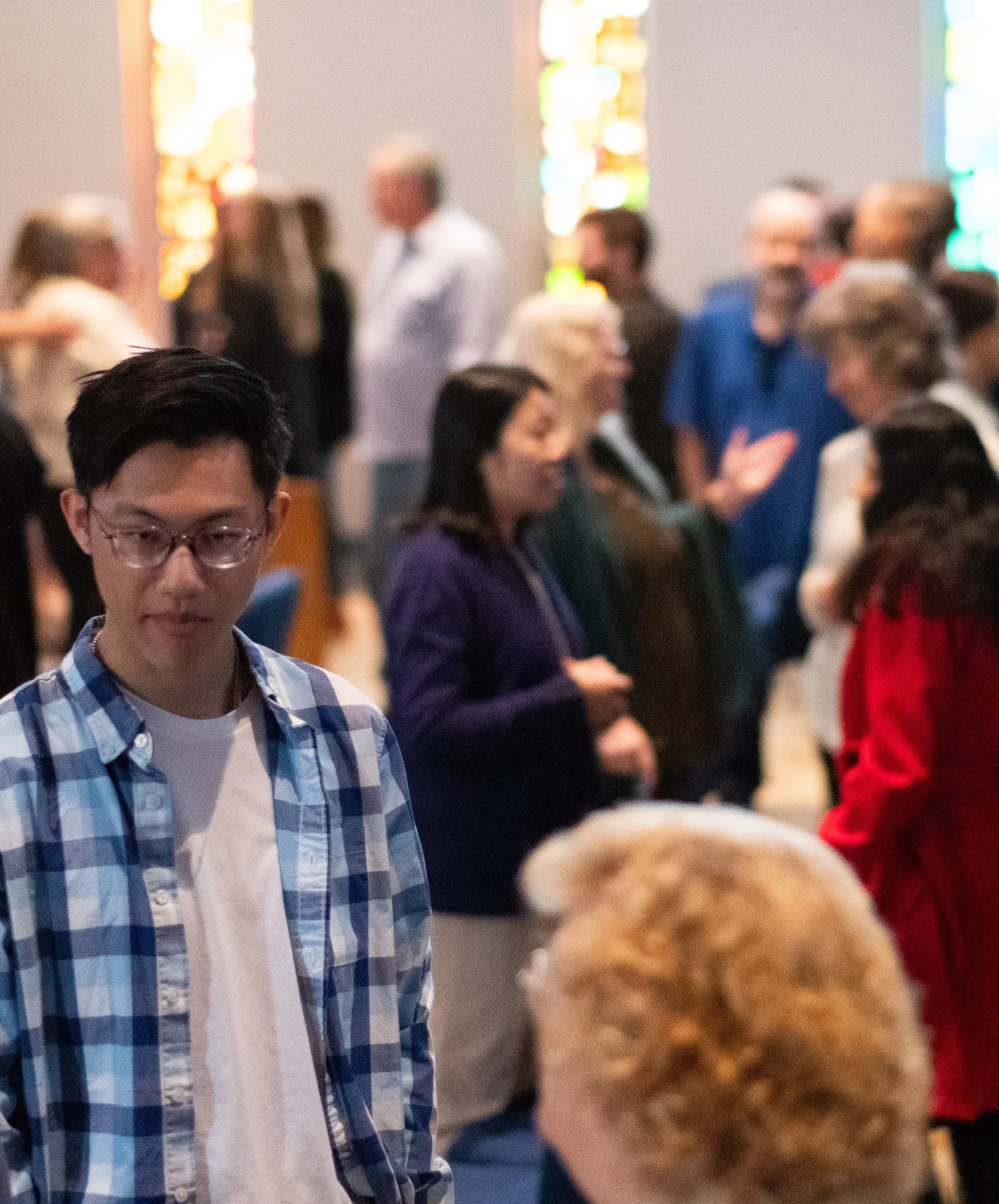 People gathering at an indoor event with colorful stained glass windows in the background, some cropped or blurred.