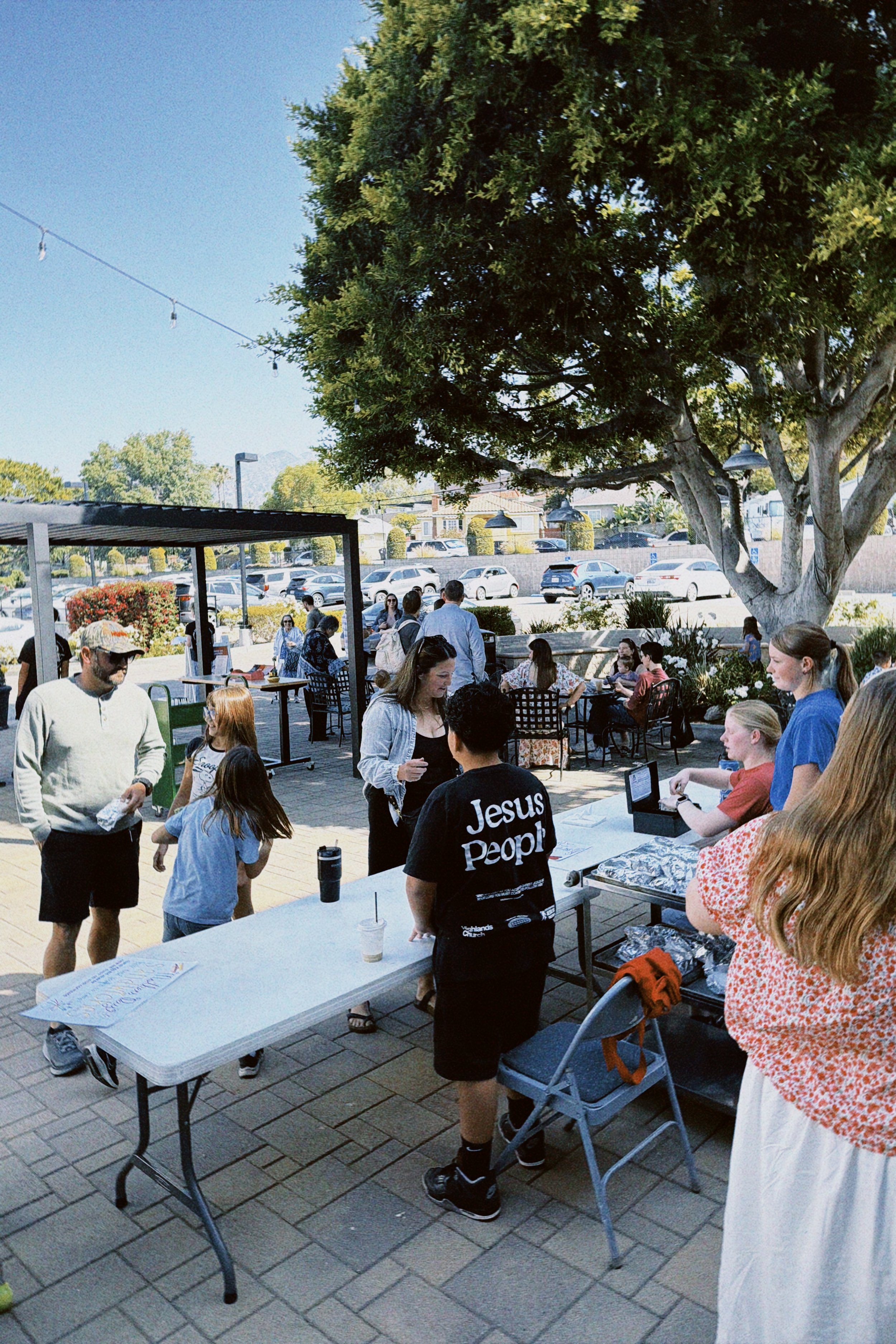 Outdoor gathering of people around a table with some food items, under a large green tree with string lights hanging above, parking lot in the background, clear sky.