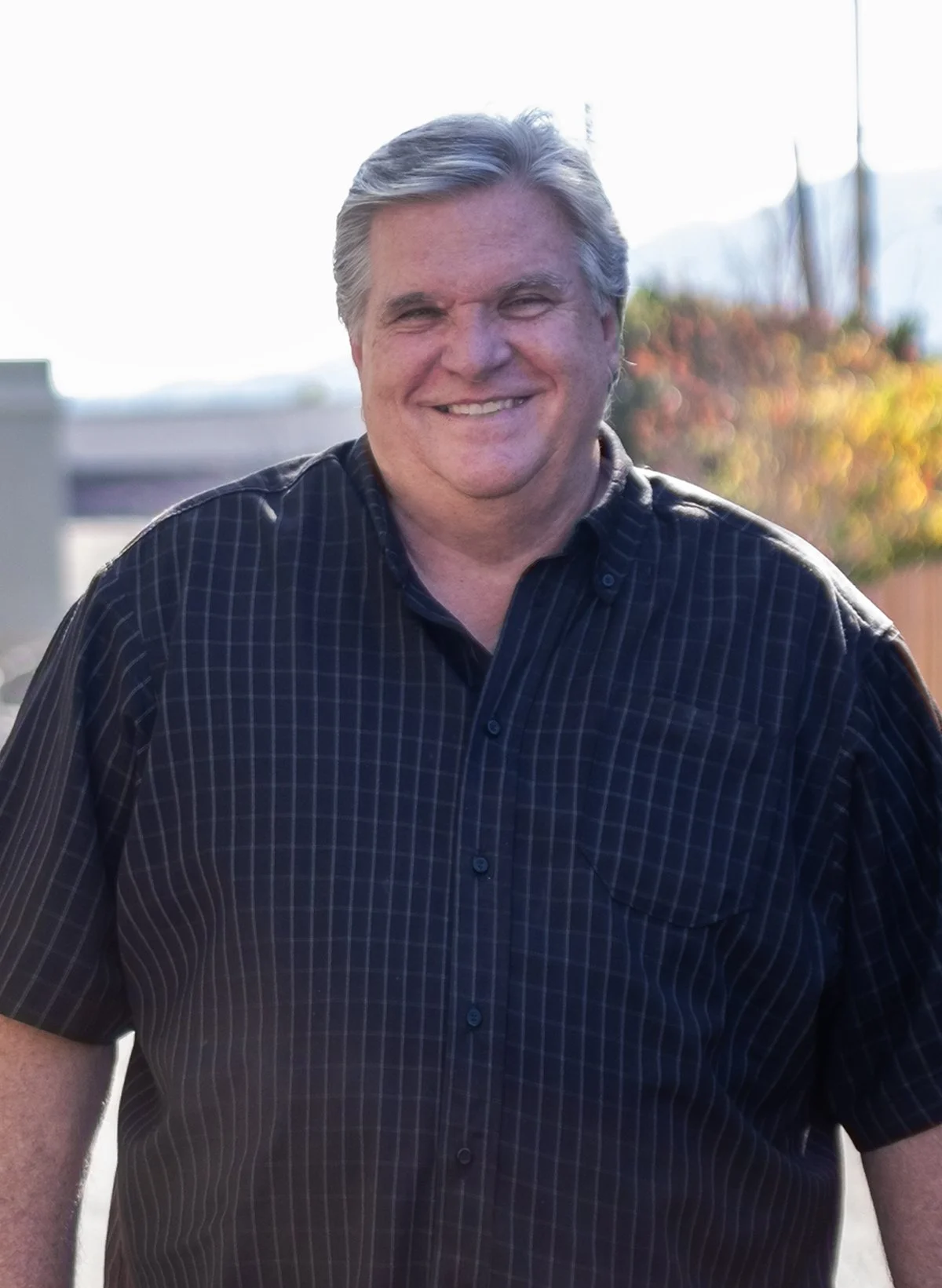 A middle-aged man with gray hair smiling outdoors, wearing a black checkered button-up shirt.