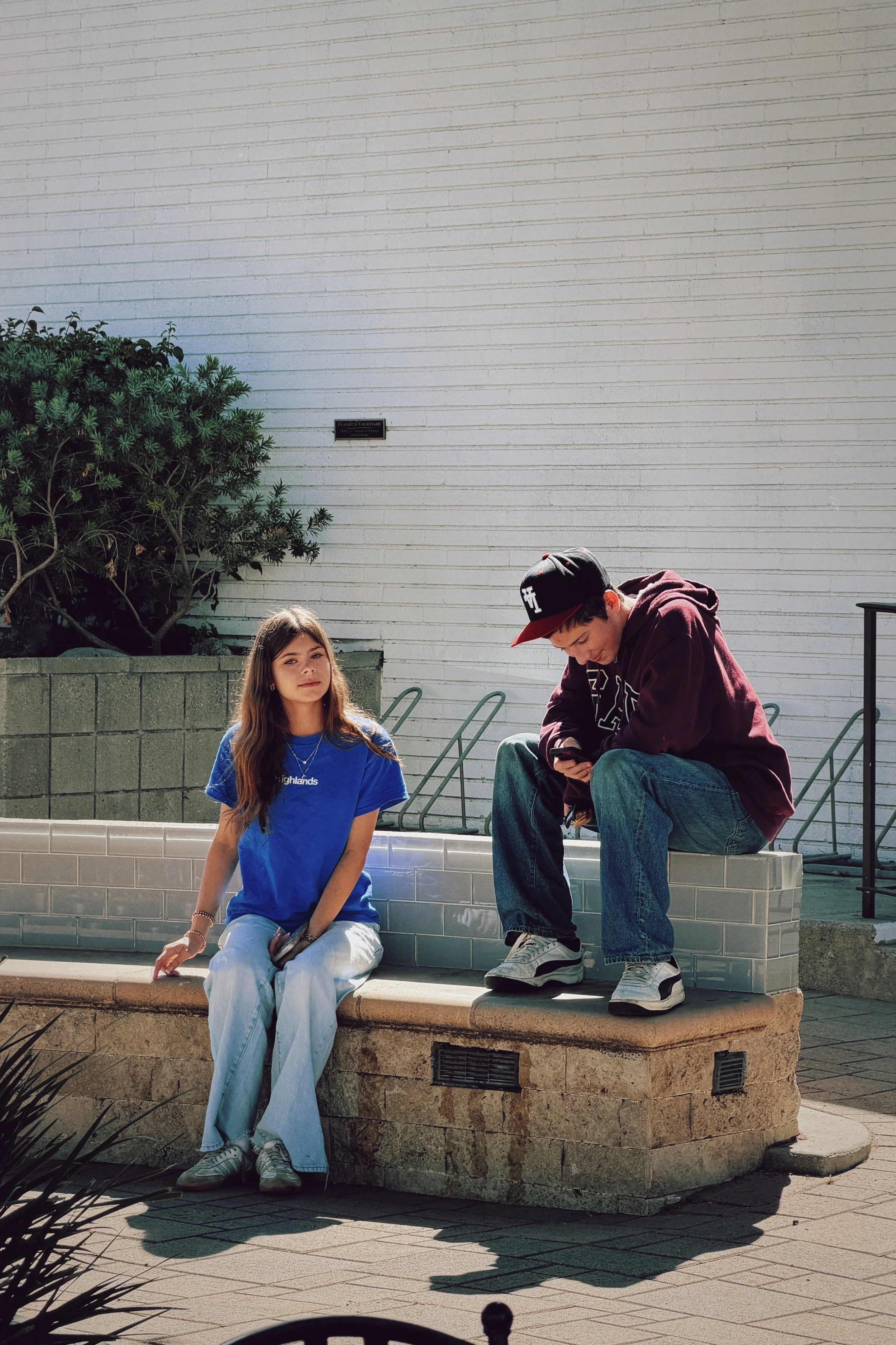 Two teenagers sitting on a stone bench in front of a white brick wall with a bush on the left. The girl on the left is wearing a blue shirt and white pants, looking at the camera, while the boy on the right is wearing a dark hoodie and jeans, looking down at his phone.