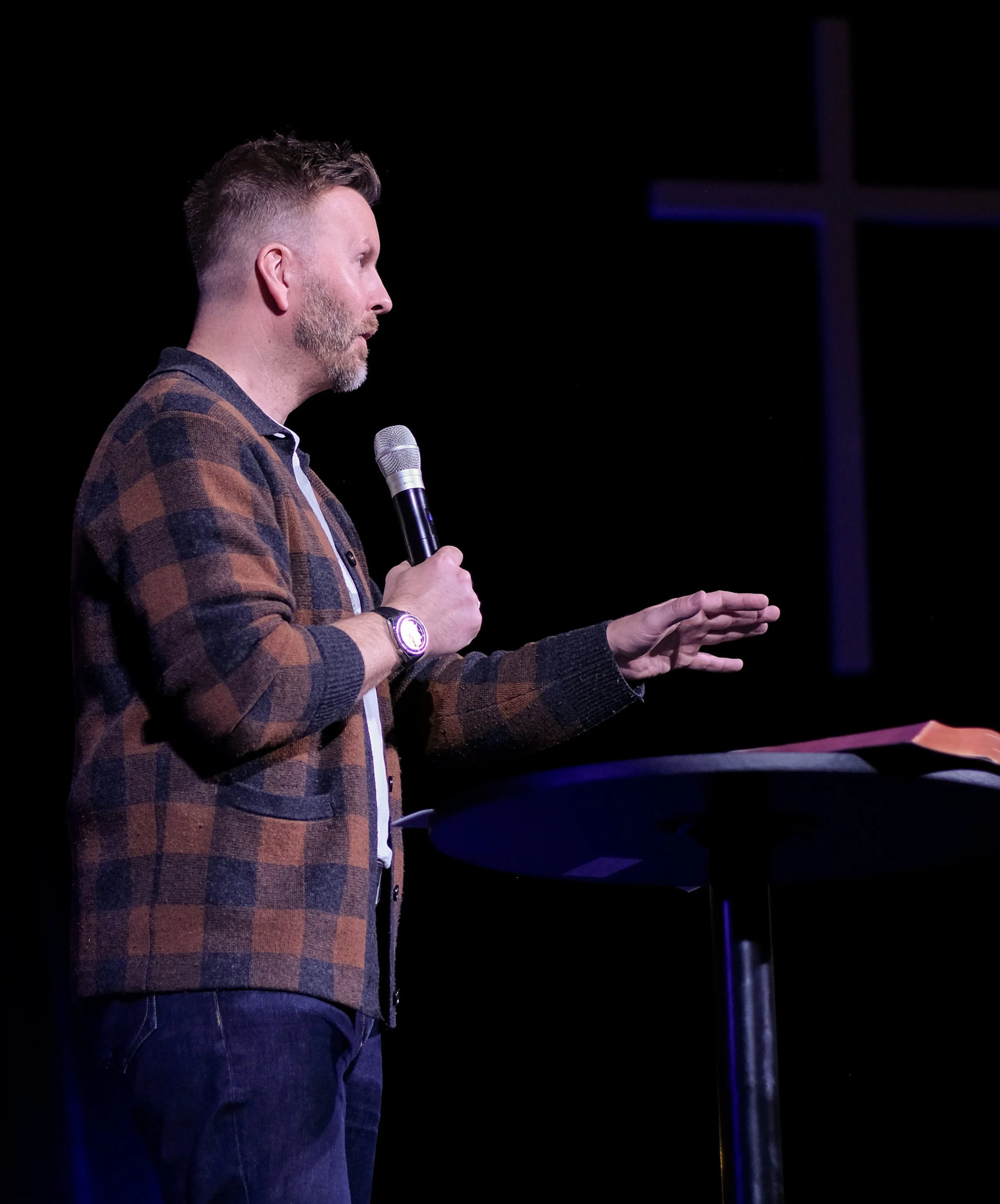 A man speaking into a microphone on stage, seen from the side, with a dark background and a cross illuminated behind him.
