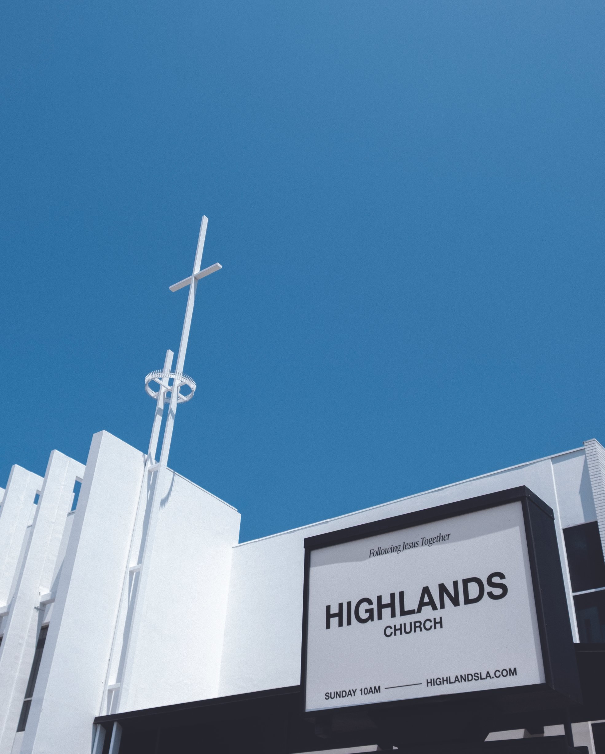 White cross on a tall pole against a clear blue sky, with a building and a sign that reads 'Following Jesus Together Highland Lakes Church' in the foreground.