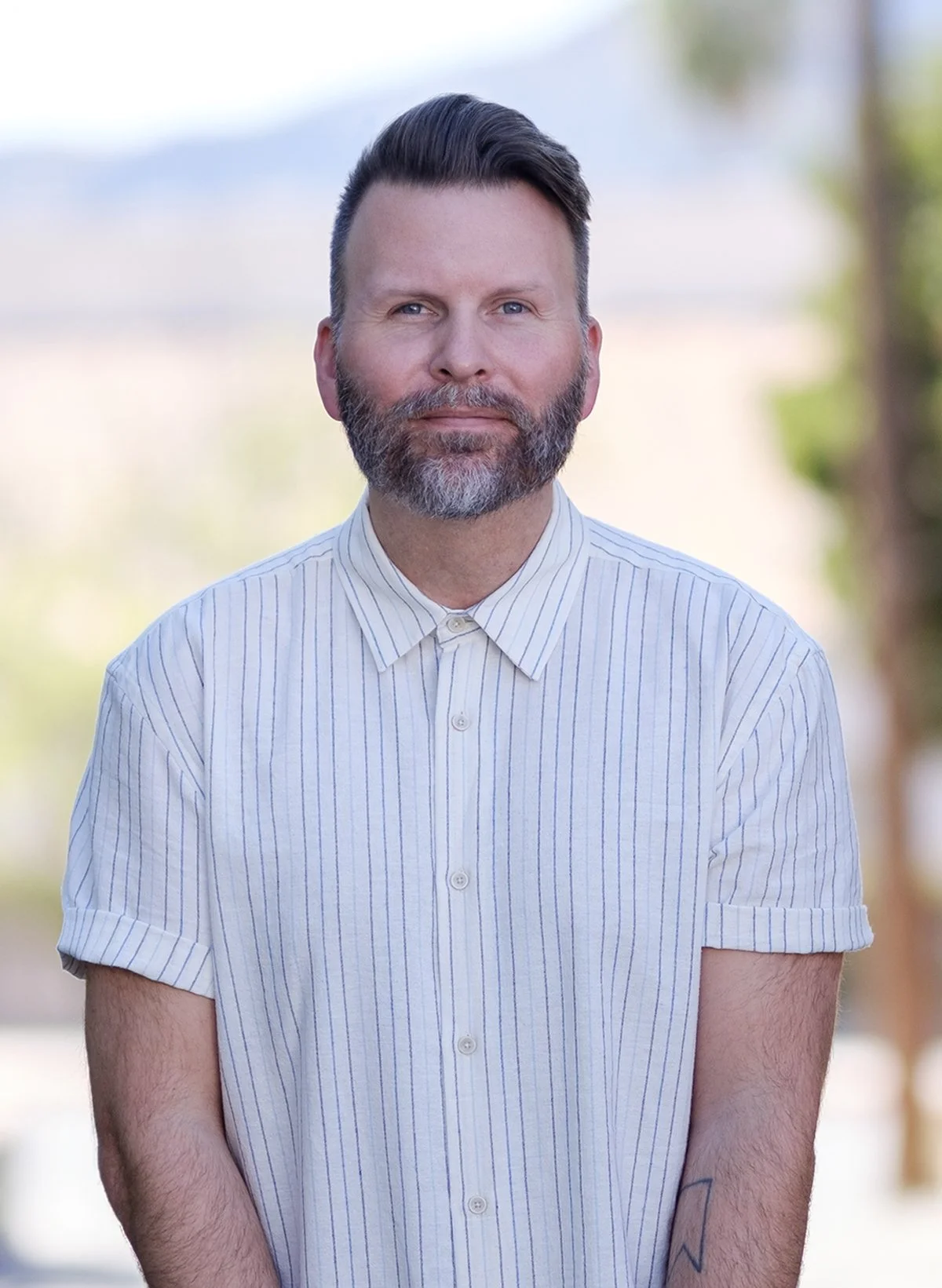 A man with a beard and short, styled hair, wearing a light striped button-up shirt, standing outdoors with blurred trees and sky in the background.