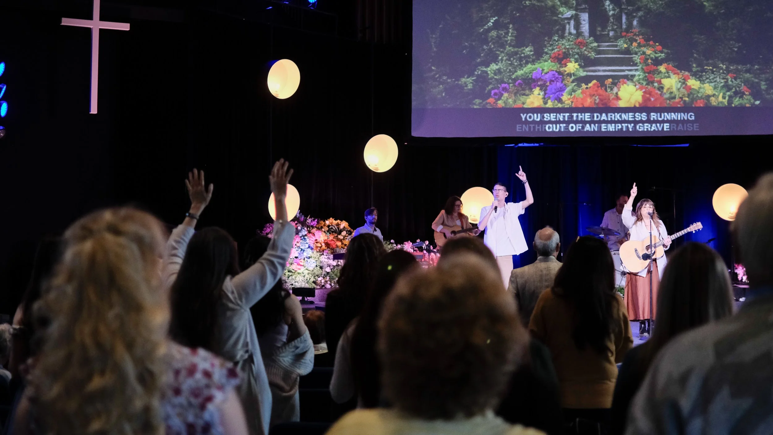 A church service or worship event with a congregation raising hands, led by musicians on stage. The stage has flowers, a cross, and a large screen displaying a garden scene with stairs and flowers, and song lyrics.