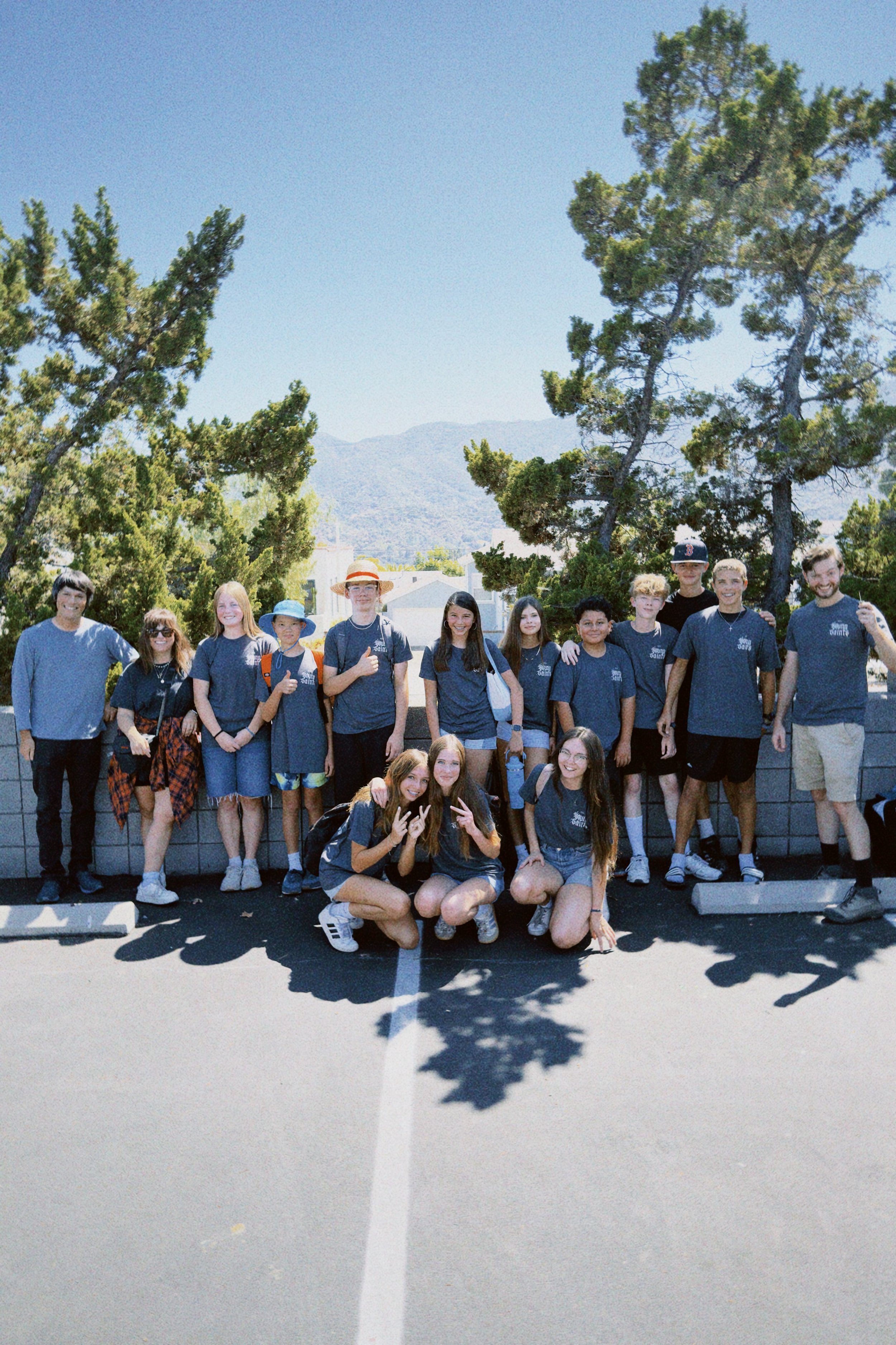 A group of twenty young people posing for a photo outdoors on a sunny day, with mountains and trees in the background, standing on a paved area with a white line and casting shadows on the ground.