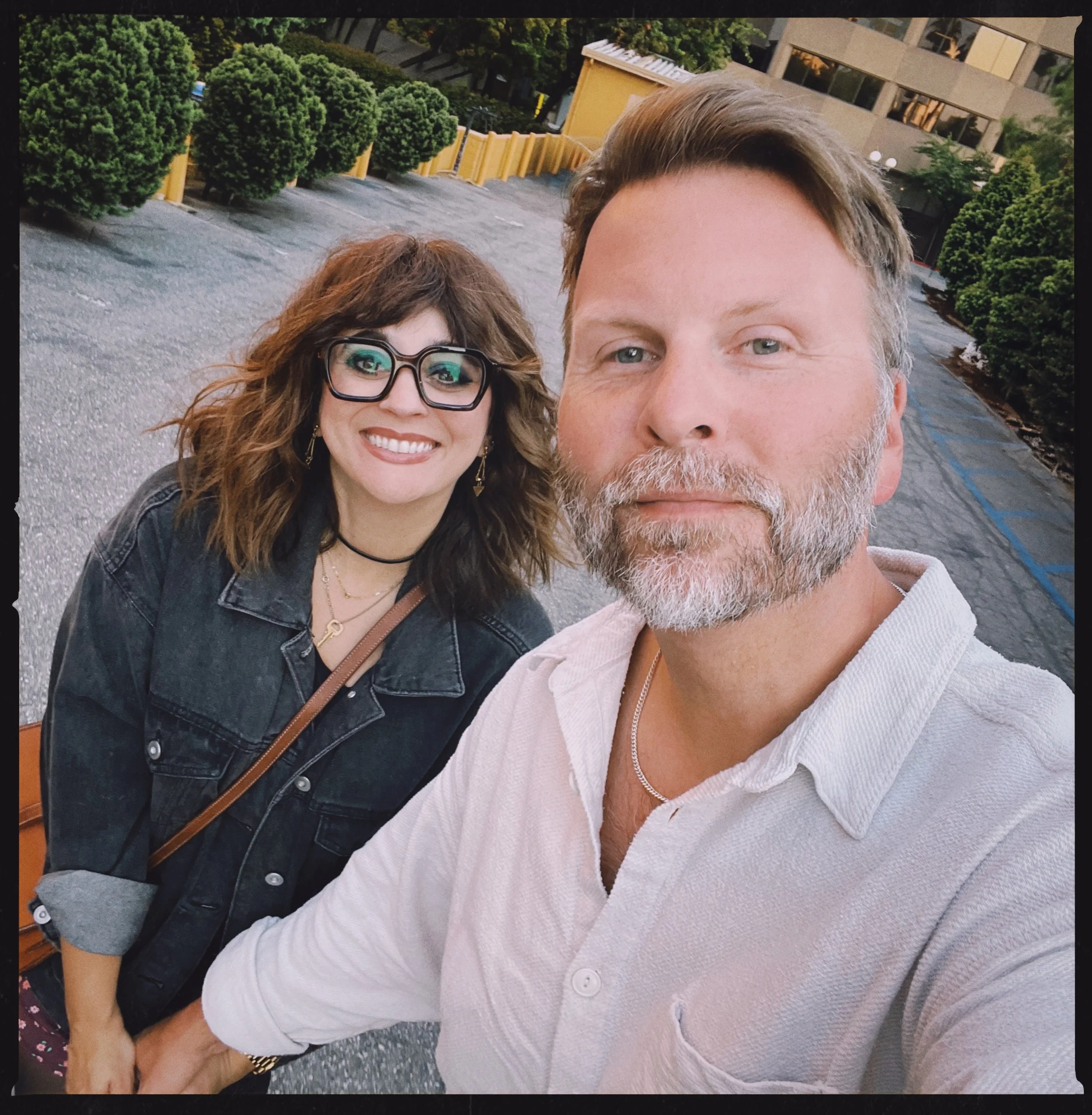 Close-up of a smiling woman with curly brown hair and glasses, and a man with light brown hair and a beard, taking a selfie outdoors near bushes and a building.