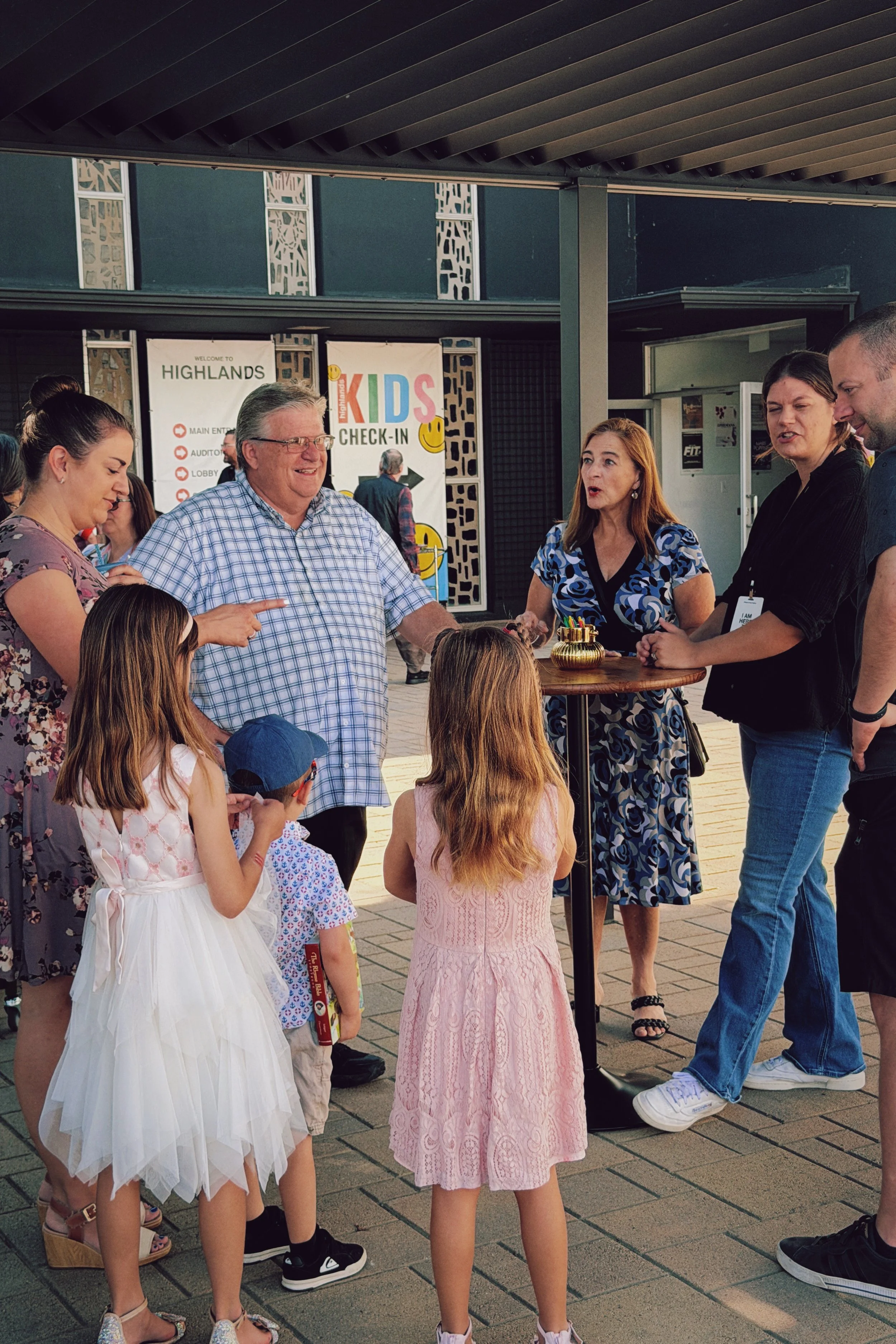 Group of children and adults gathered outdoors in conversation, with signs in the background indicating a Highlands event and kids check-in.