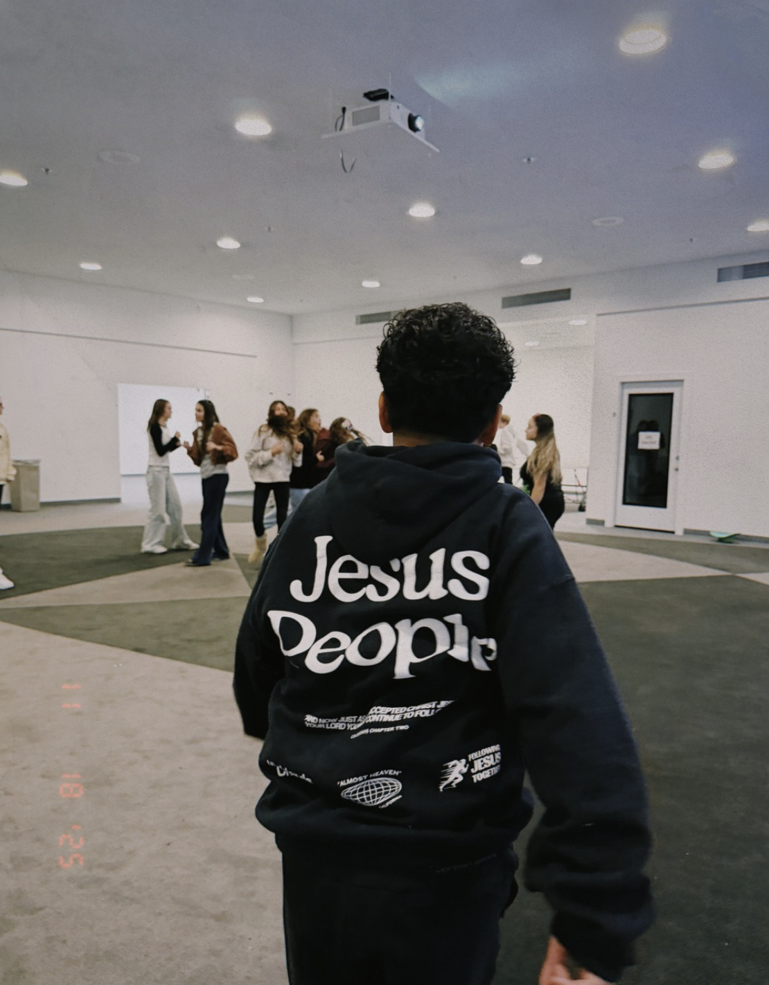 A person wearing a black hoodie with the words 'Jesus People' stands in the foreground, facing a group of people in a large indoor space, possibly a church or community center, with white walls and a projector on the ceiling.