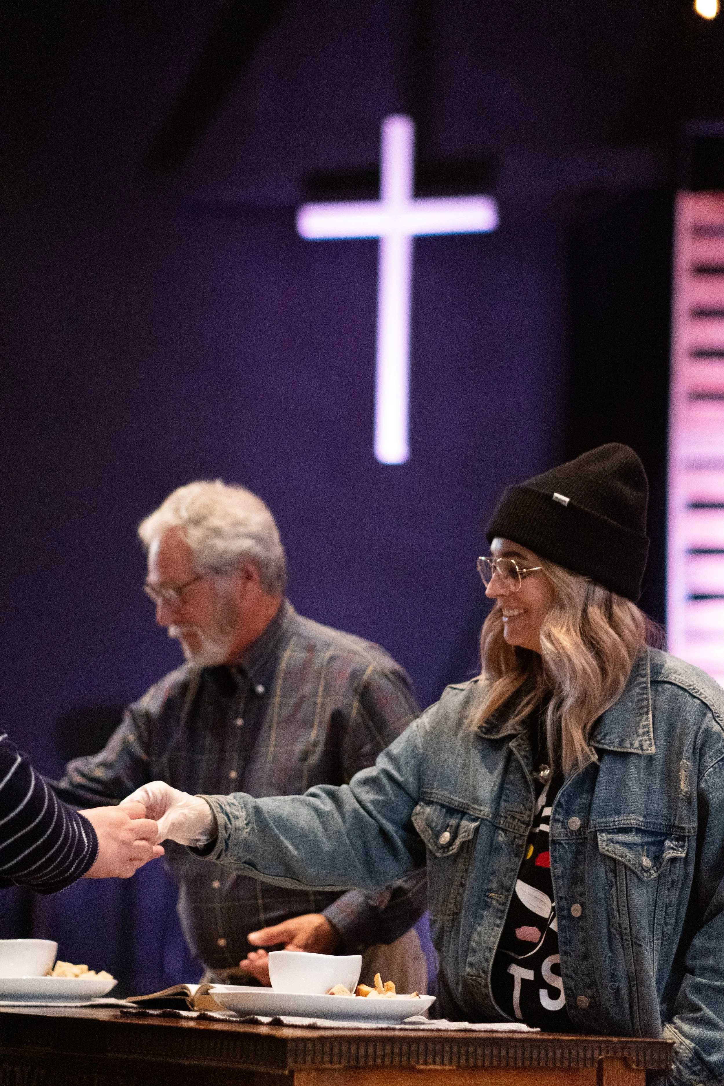 People shaking hands across a table with bowls of food in a dimly lit room with a illuminated cross in the background.