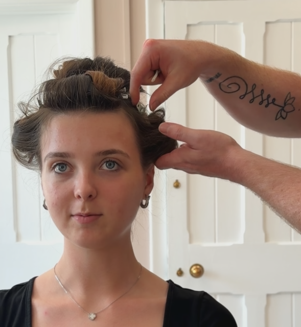 A woman is getting her hair styled or prepared, with a person working on her hair in a room with white paneled doors.