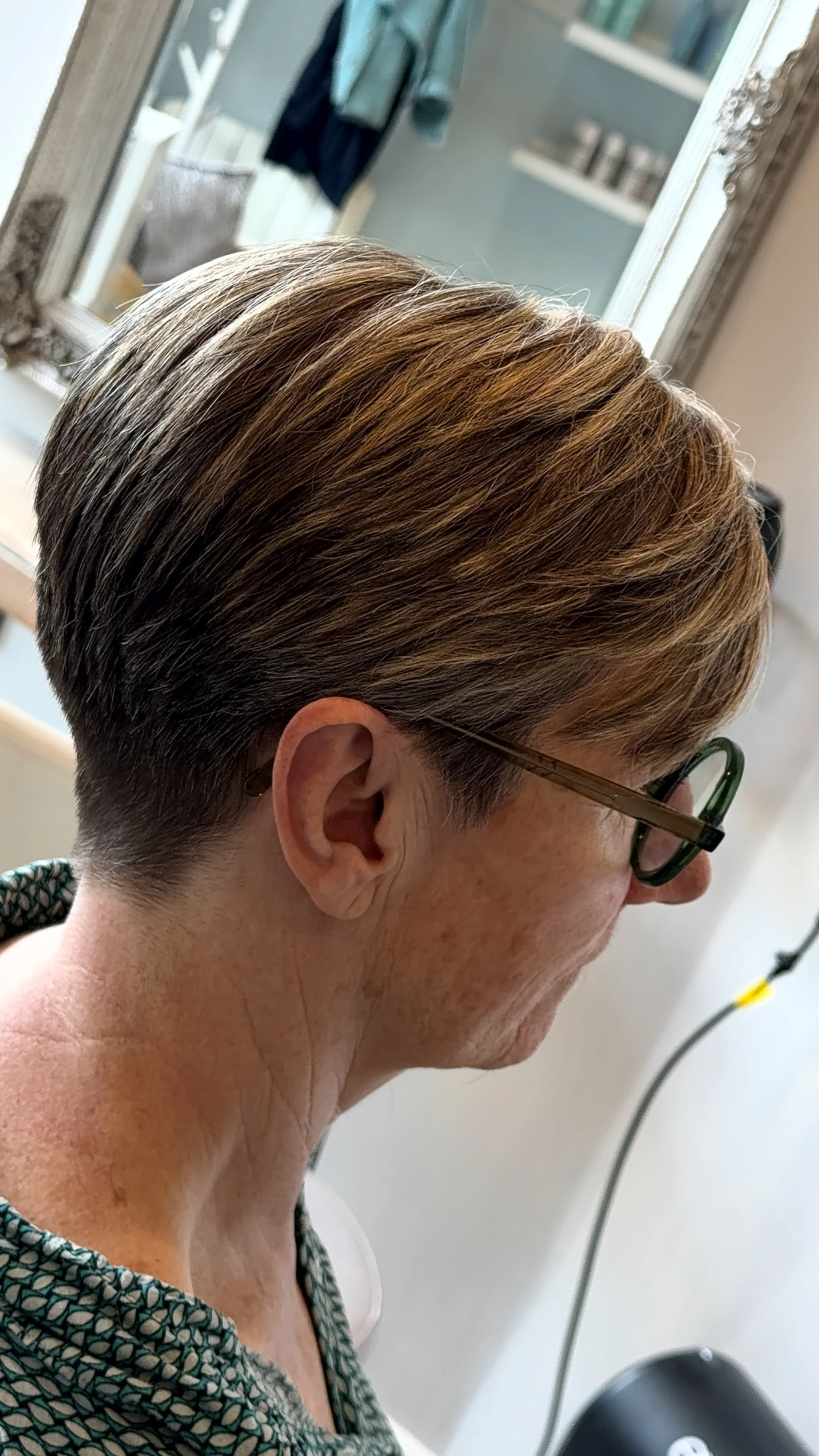 Side view of a woman with short, styled hair, wearing glasses, in a salon or bathroom setting.