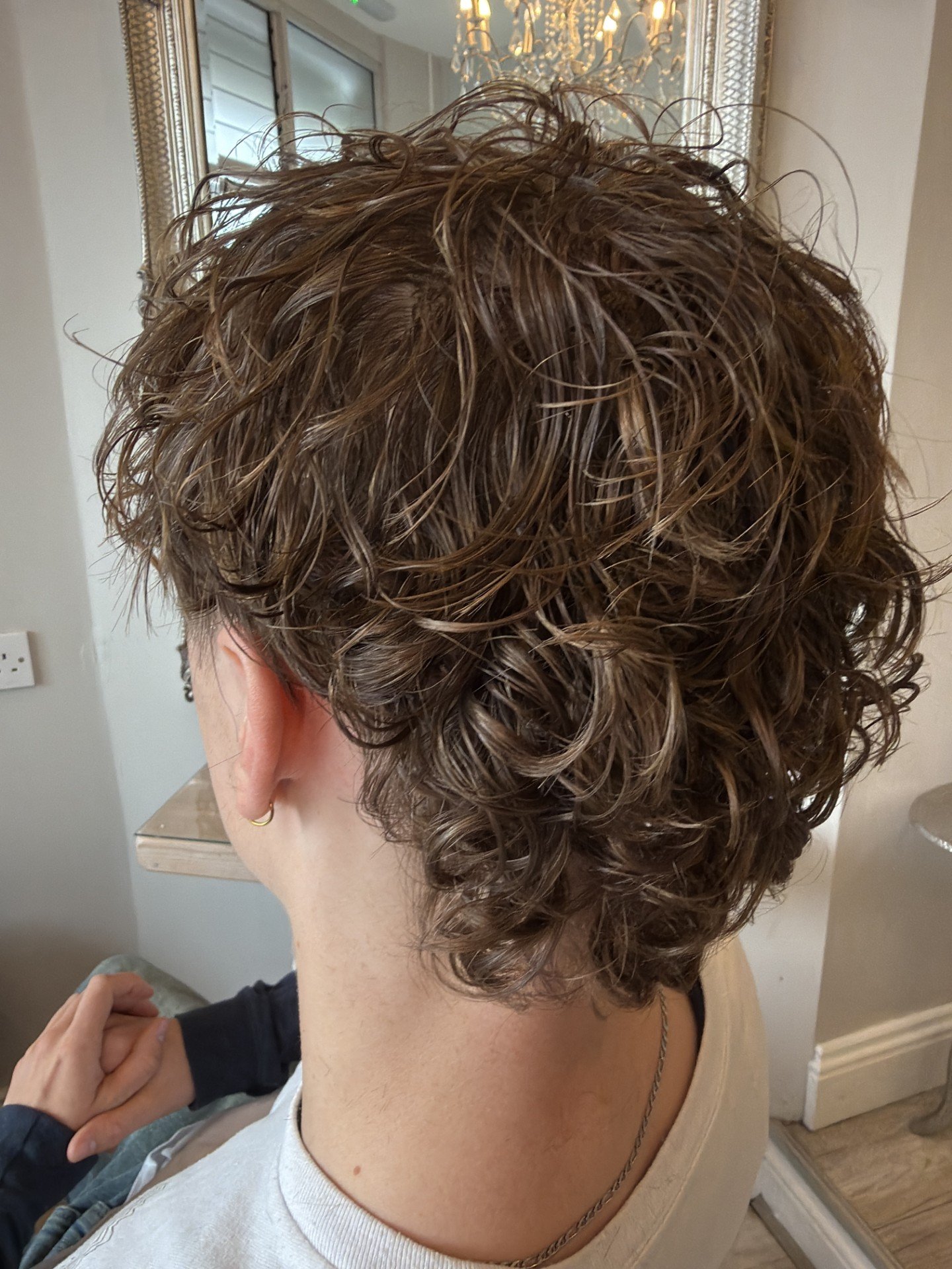 Back view of a person with short, curly, light brown hair. The person is wearing a cream-colored top and has earrings and a necklace. They are sitting in a room, likely a living space with a mirror and lighting fixture visible in the background.