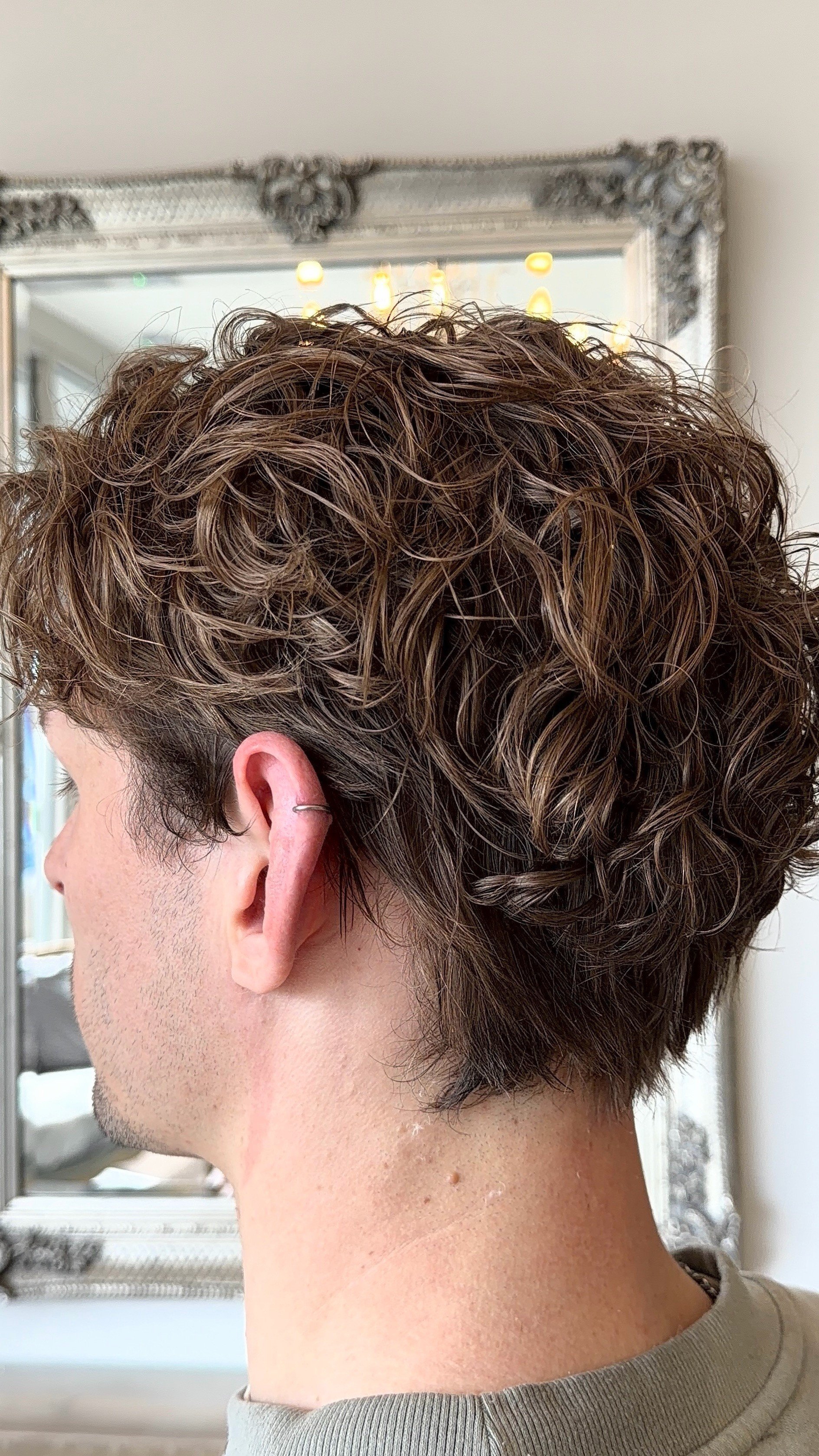 A person with short, curly, brown hair with highlights, seen from the side in a salon or mirror setting.