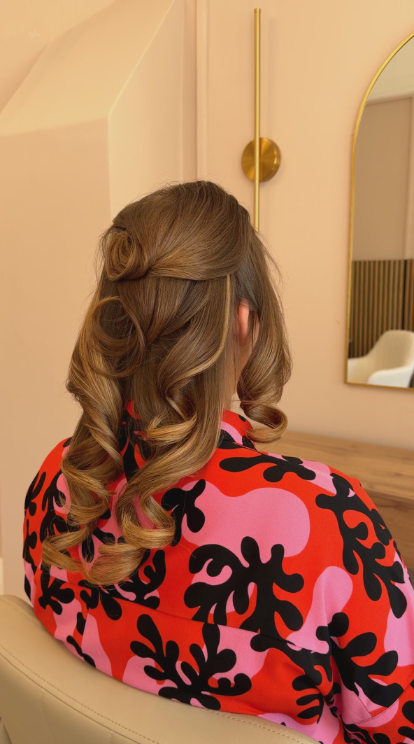 A woman with styled, curled brown hair sitting in a room with beige walls, a mirror, a chair, and modern gold wall decor.