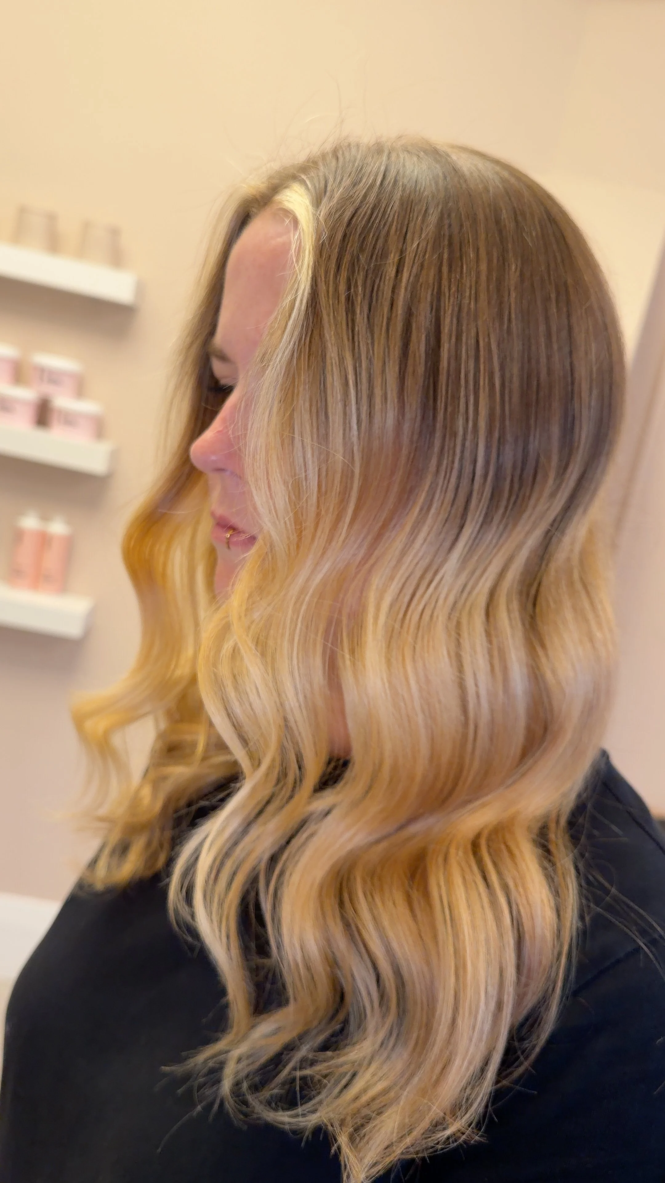 Side profile of a woman with long, wavy blonde hair, wearing a black top, standing in a room with beige walls and shelves with pink products in the background.