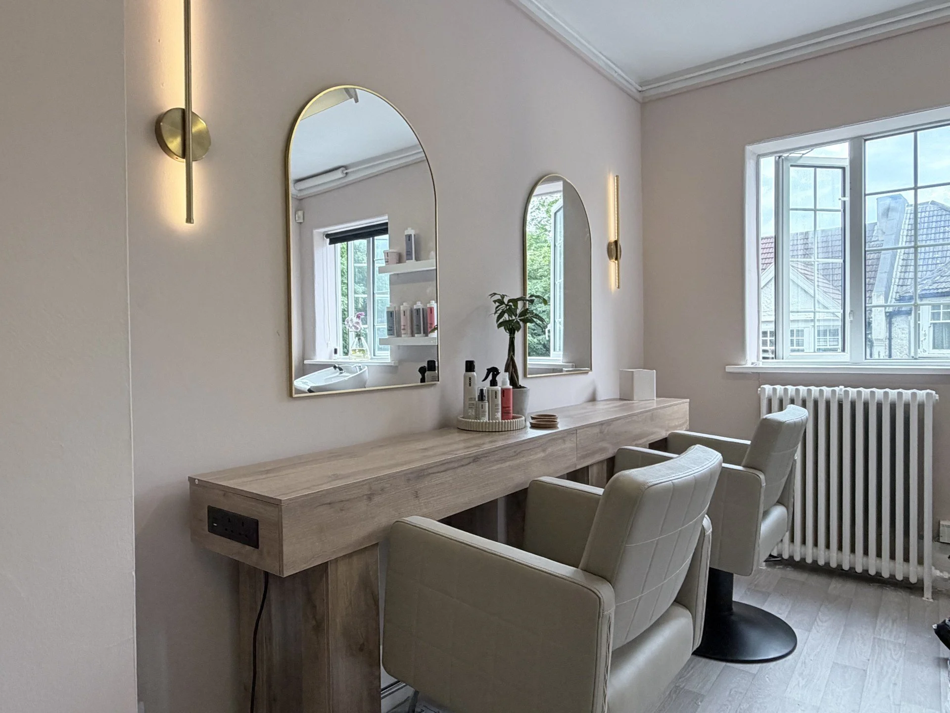 A salon or hair studio with a wooden countertop, two white salon chairs, and large windows with buildings outside. The wall has two oval mirrors and modern wall lights.