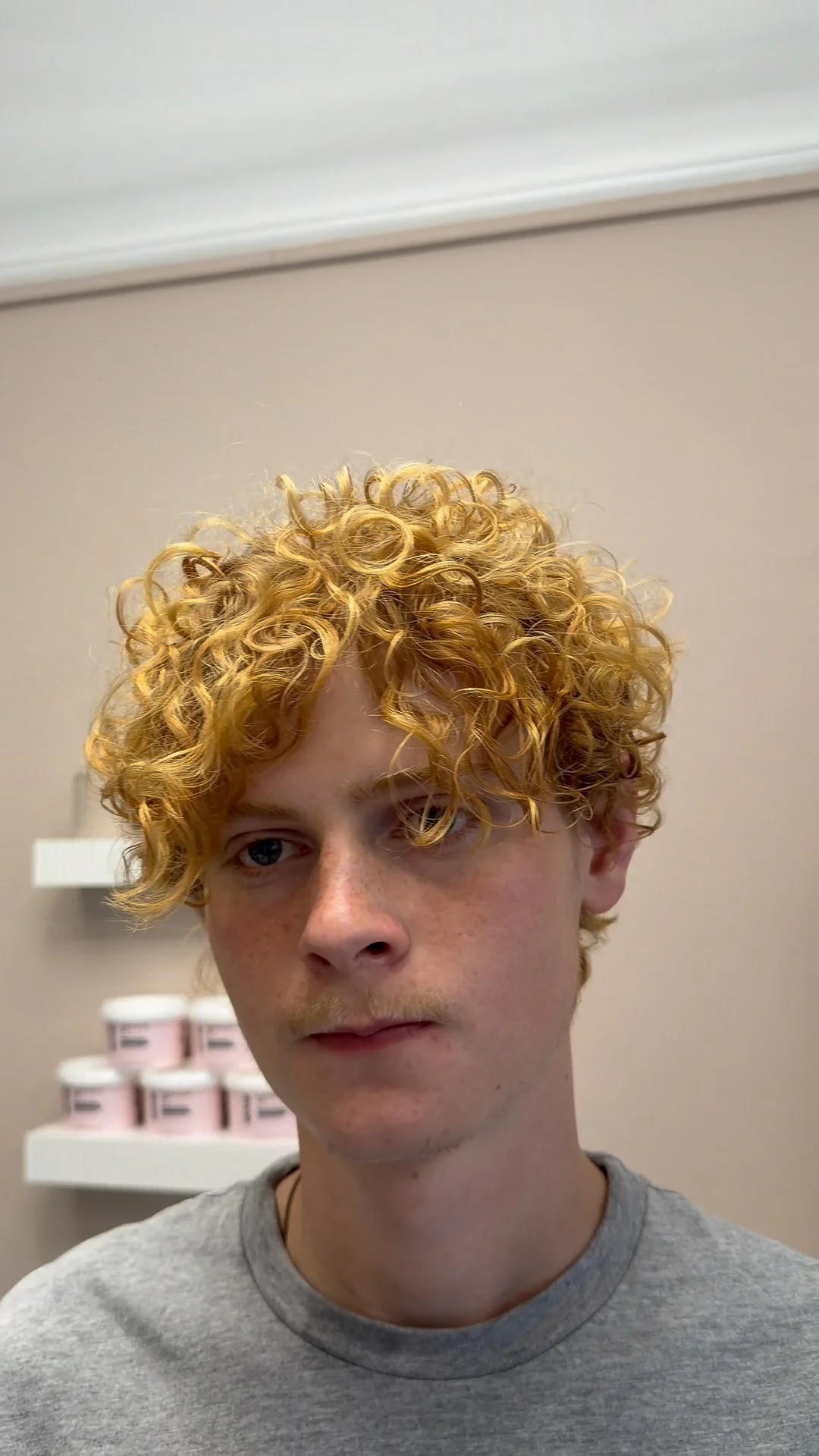A young man with curly blonde hair and light skin looking to the side, wearing a gray t-shirt, in a room with shelves of pink and white containers in the background.