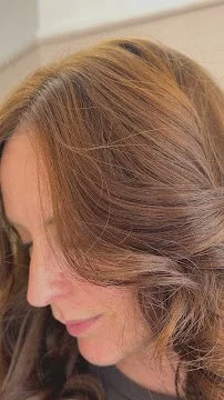 Close-up of a woman's head showing her brown, wavy hair and a side profile of her face with a relaxed expression.