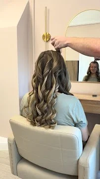 A person with long, curled hair sitting in a salon chair, while another person's hand is adjusting a lock of hair near a mirror.