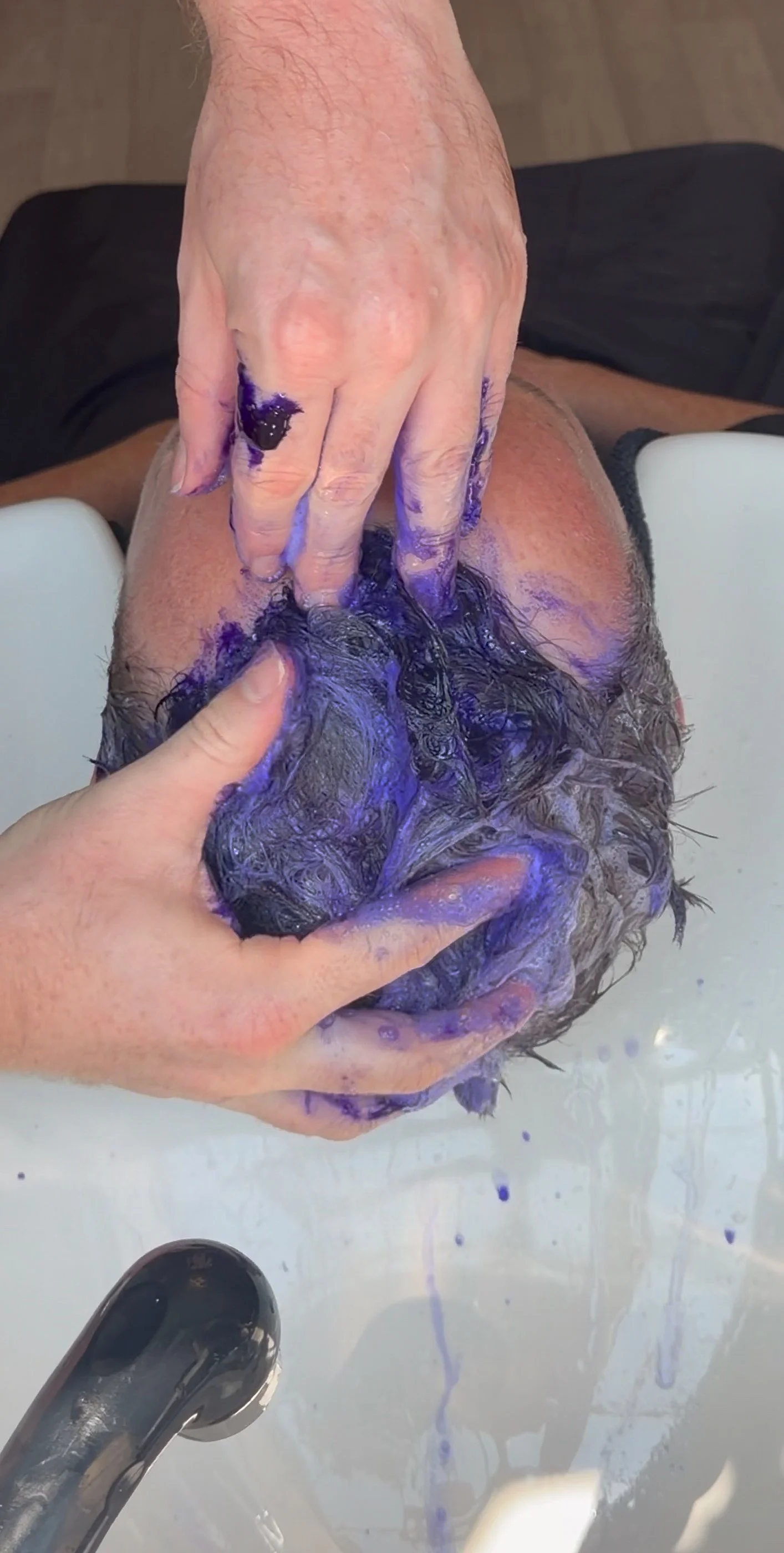 Person washing hair in a sink with purple shampoo, hands lathering and rinsing hair.