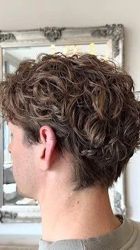 Back view of a person with short, curly brown hair, standing in front of a mirror in a salon or room with ornate framing.