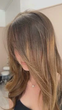 A person with long, layered brown hair covering their face, wearing a black top and a necklace, in an indoor setting.