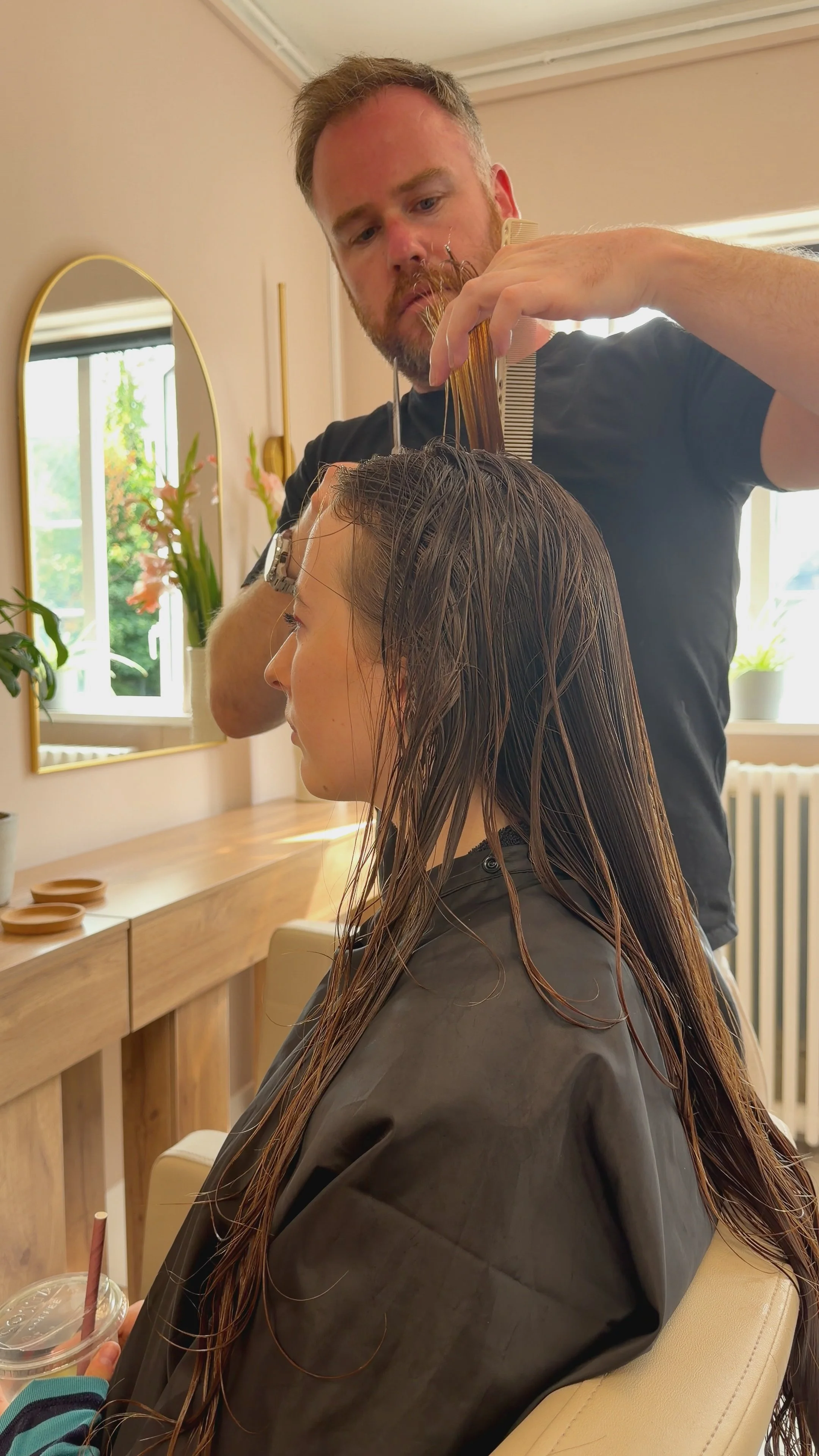 A hairstylist cutting a woman's wet hair in a salon, with a mirror and plants in the background.